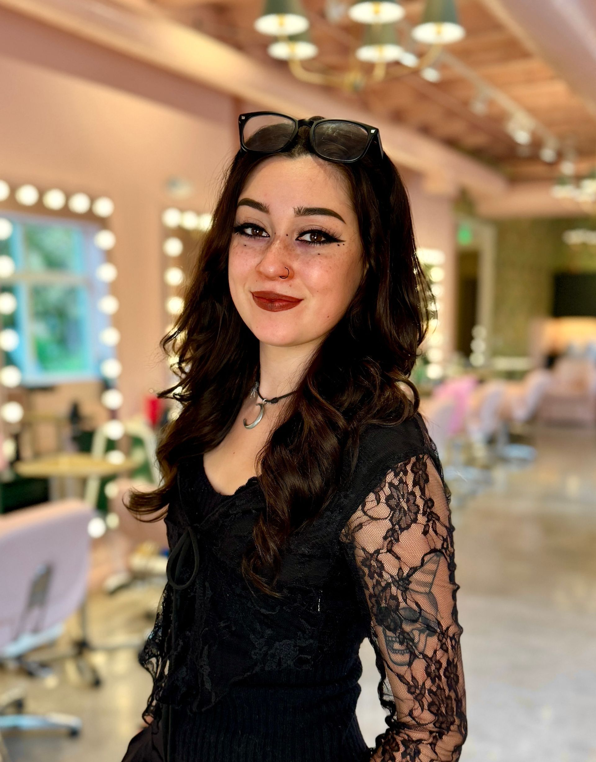 A smiling person with long brown hair, wearing a black and white dress with decorative buckles, standing in a salon.