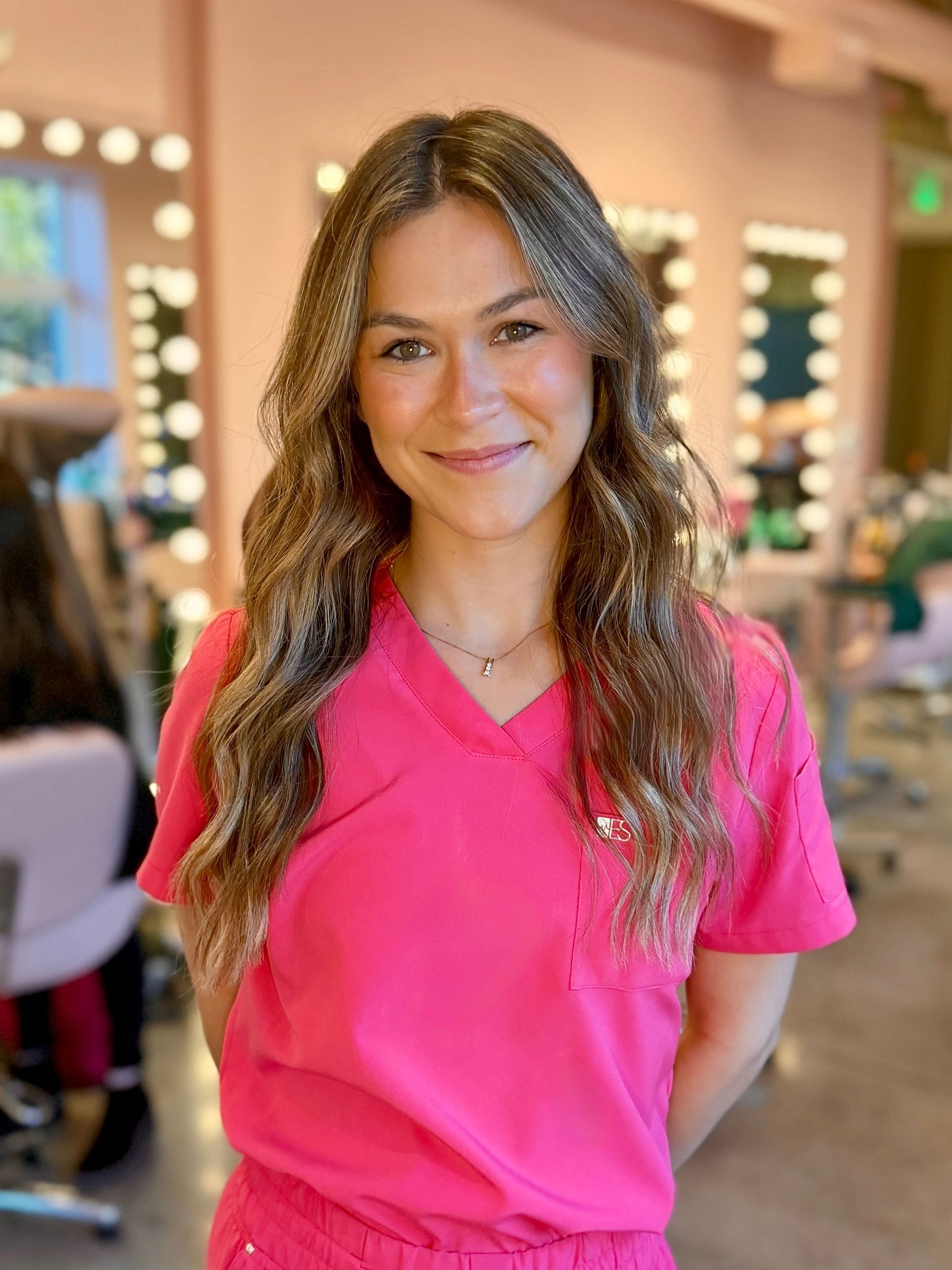 Woman smiles at the camera, wearing a pink sleeveless top; neutral background.