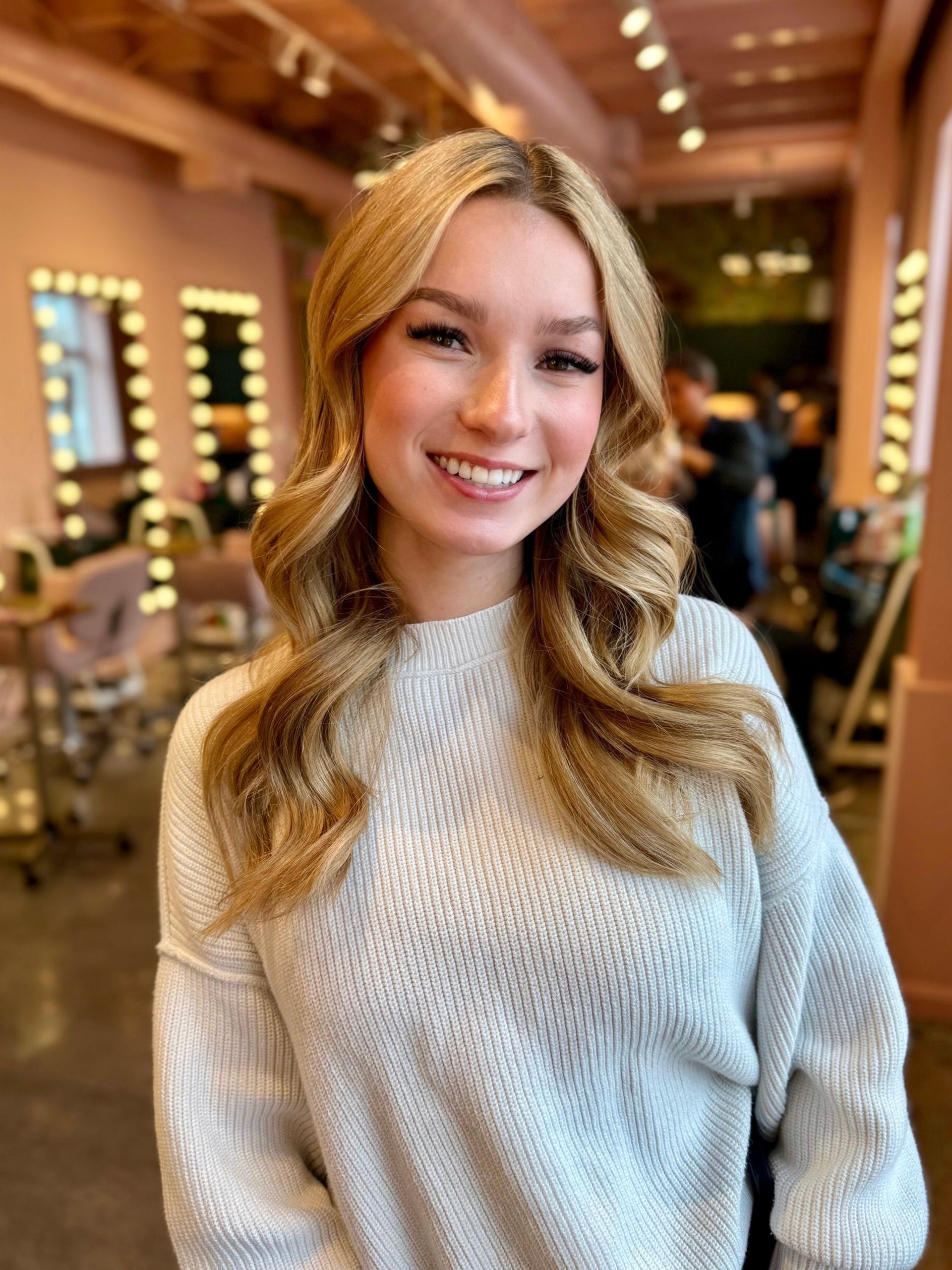 A smiling person with long blonde hair, wearing a white textured sweater, standing in a brightly lit salon.