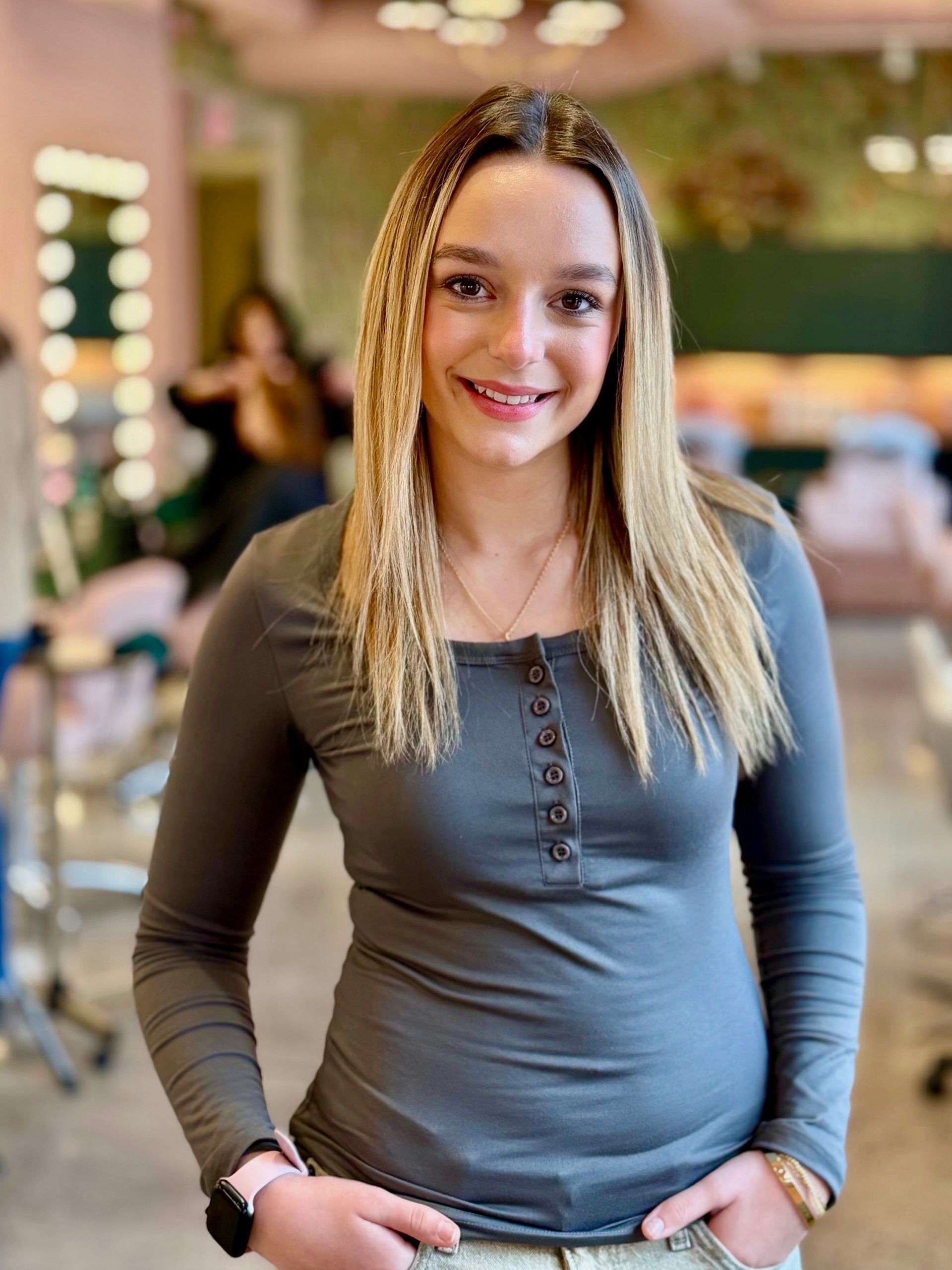 A smiling person with crimped blonde hair wearing a grey long-sleeved henley shirt in a salon setting.