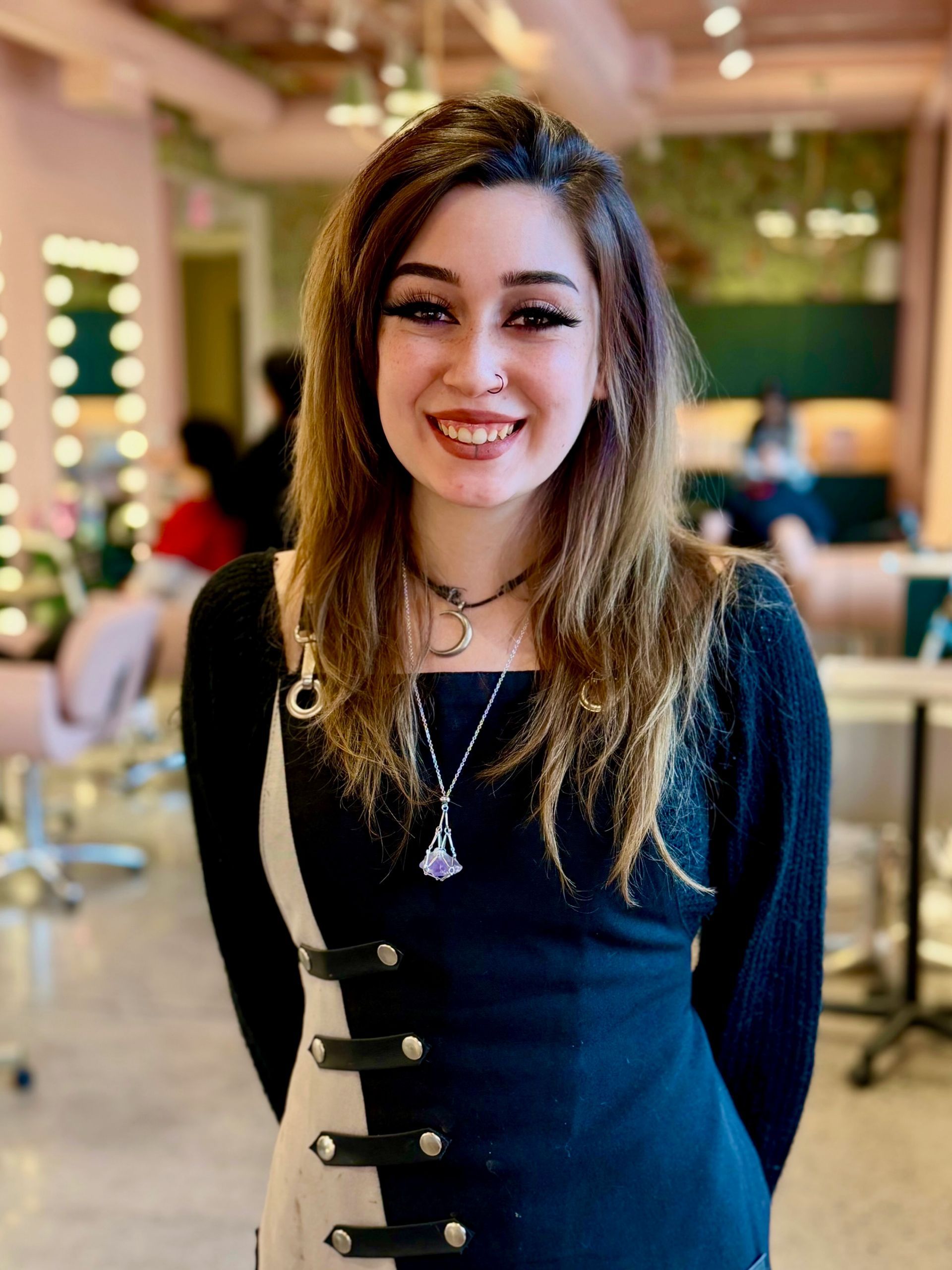 A smiling person with long brown hair, wearing a black and white dress with decorative buckles, standing in a salon.