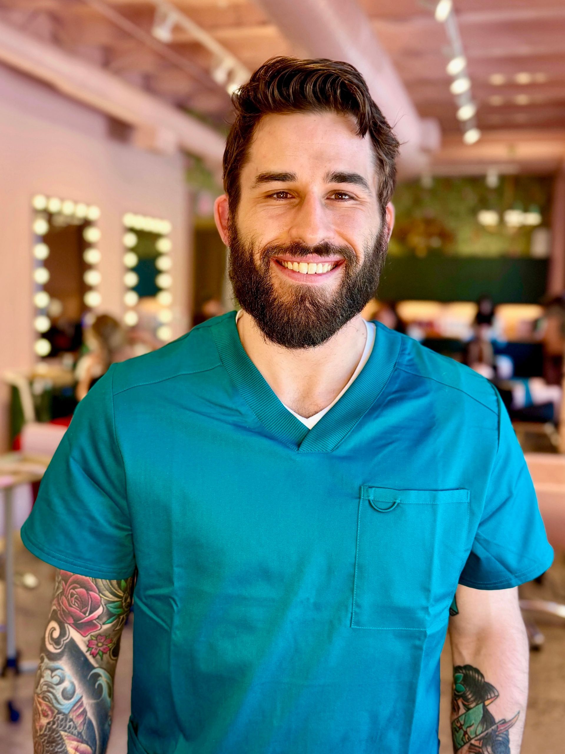 Smiling bearded man in teal scrubs standing indoors with tattooed arms