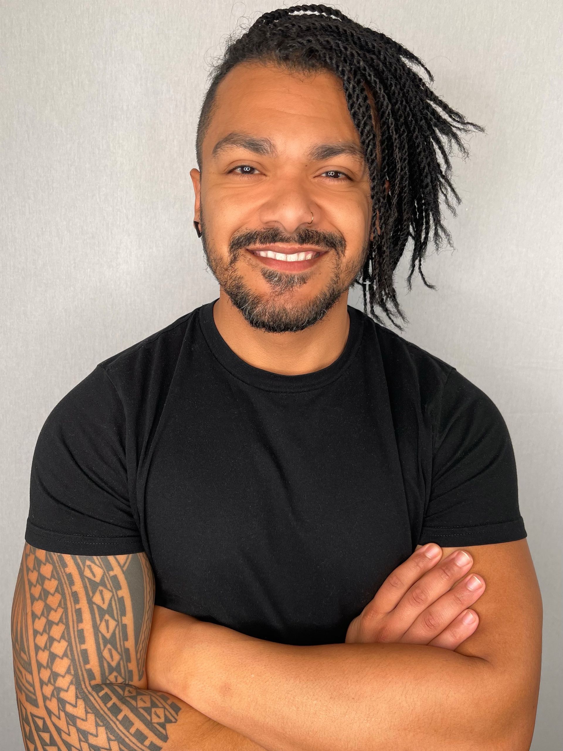 Smiling man in black T-shirt with tattooed arm, standing with crossed arms against a light wall