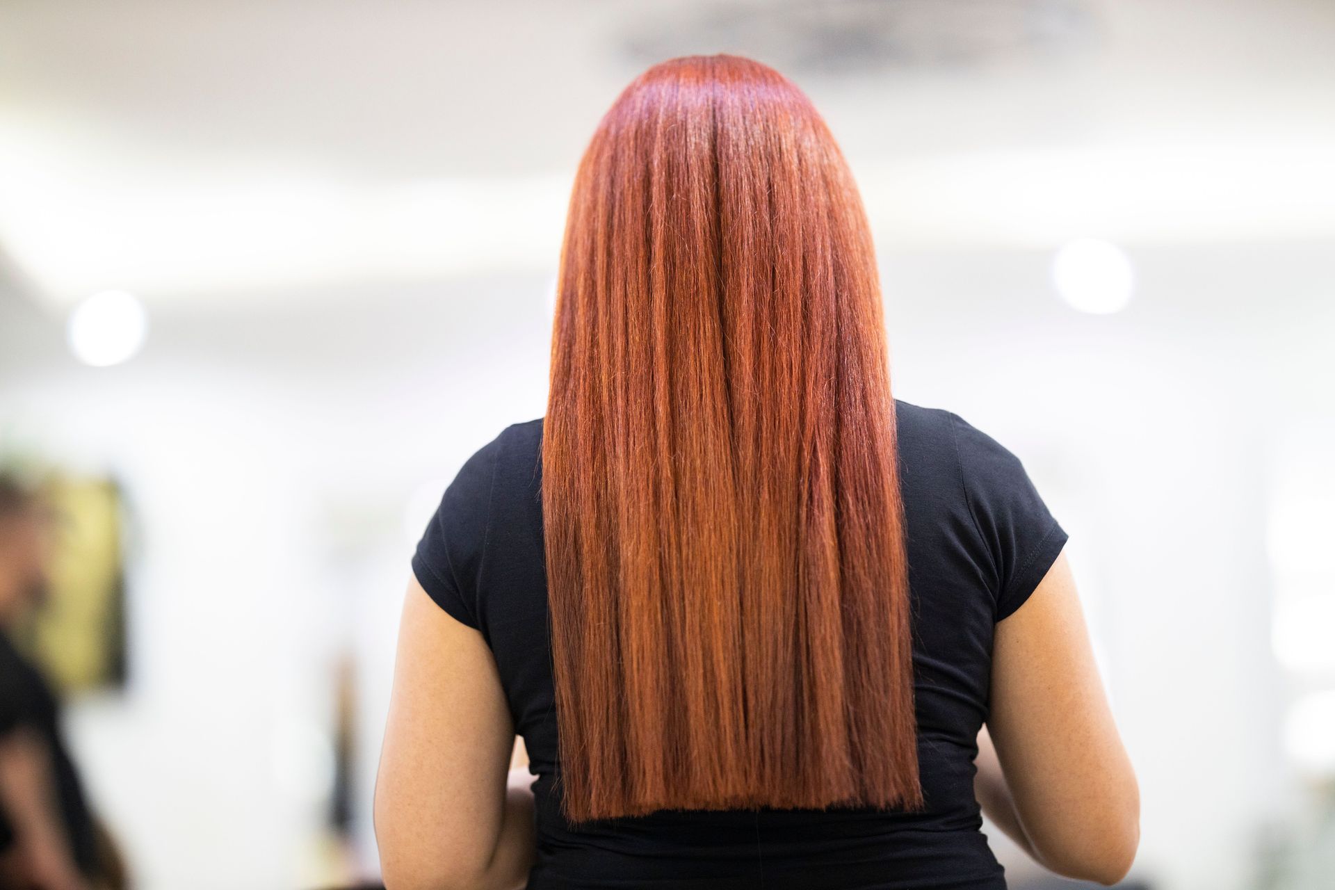 Person with long straight red hair seen from behind in a black shirt, standing indoors.