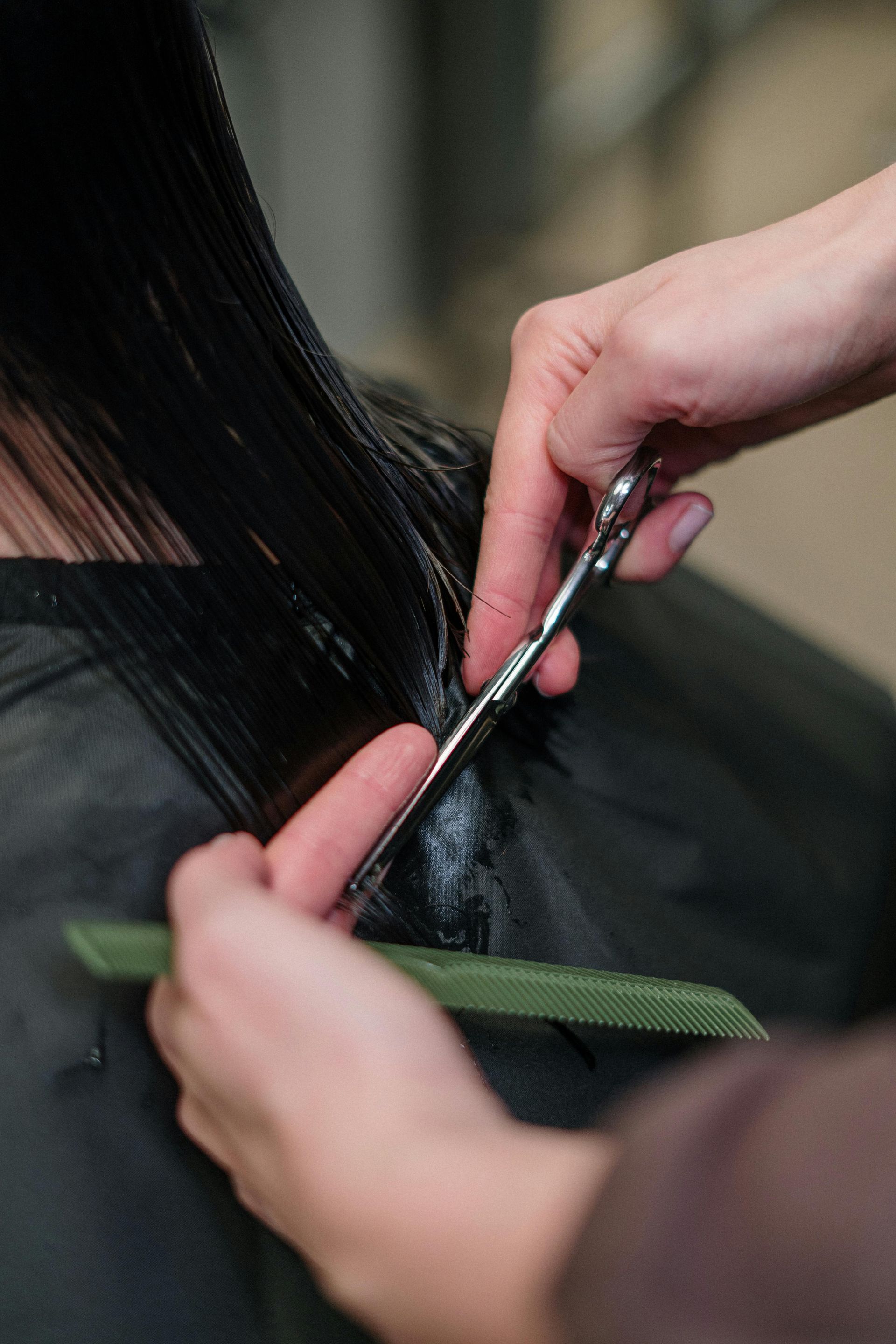 Close-up of a stylist’s hands using silver scissors and a green comb to trim straight, dark hair.