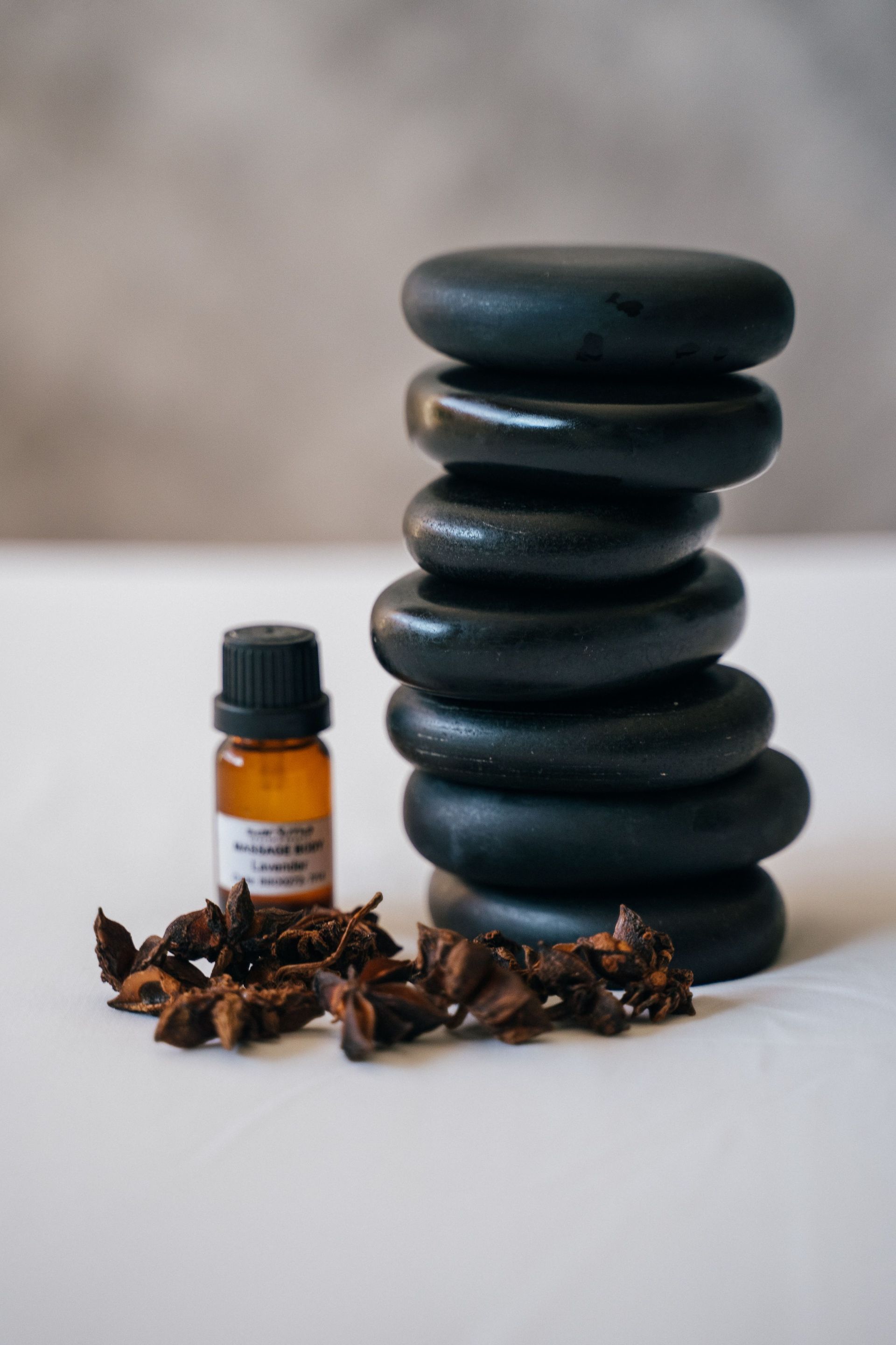 Black spa stones stacked beside a small amber essential oil bottle with dried cloves on a white surface