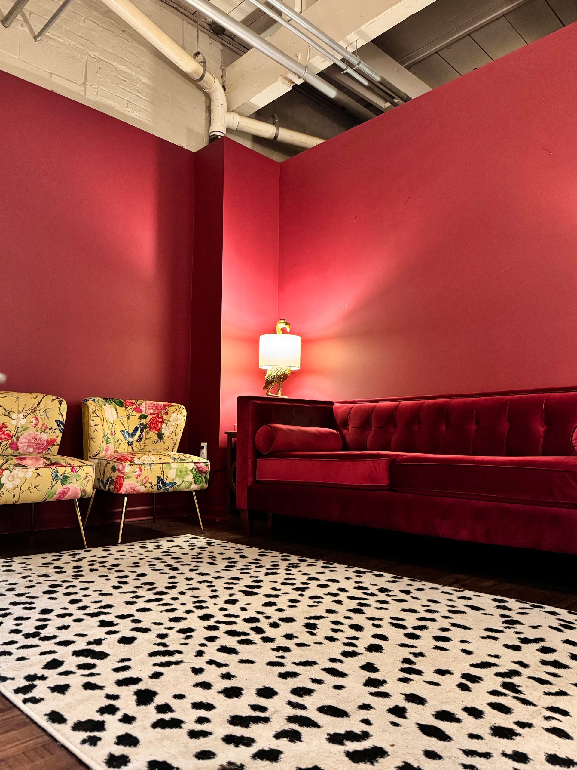 Colorful lounge with red walls, floral chairs, a velvet sofa, and a spotted floor lamp