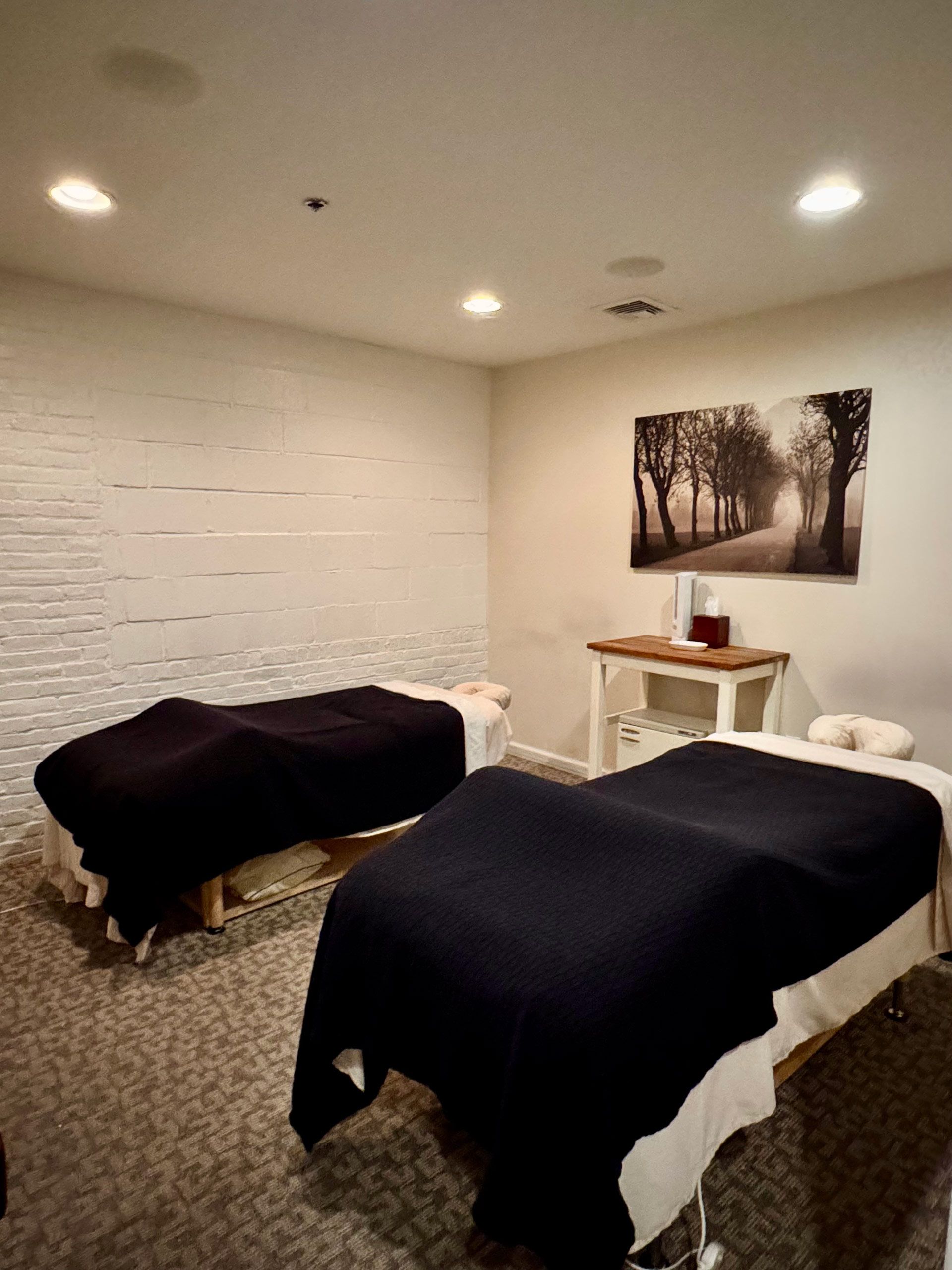 Massage room with two covered tables, soft lighting, and a wall photo above a small shelf.