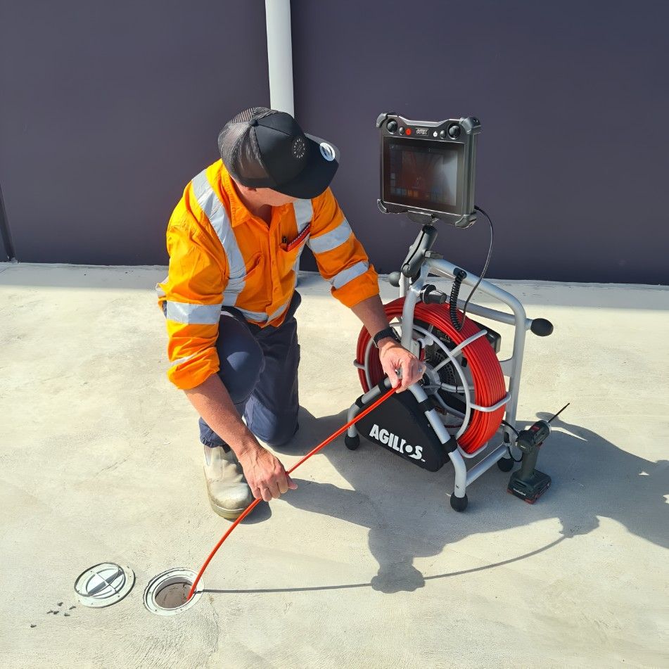A Man in an Orange Shirt is Kneeling Down Next to a Drain Camera — Dowds Pipe and Cable Locating in Farmborough Heights, NSW