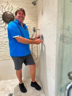 Man in blue shirt smiling in a shower, holding the controls by the tiled wall.
