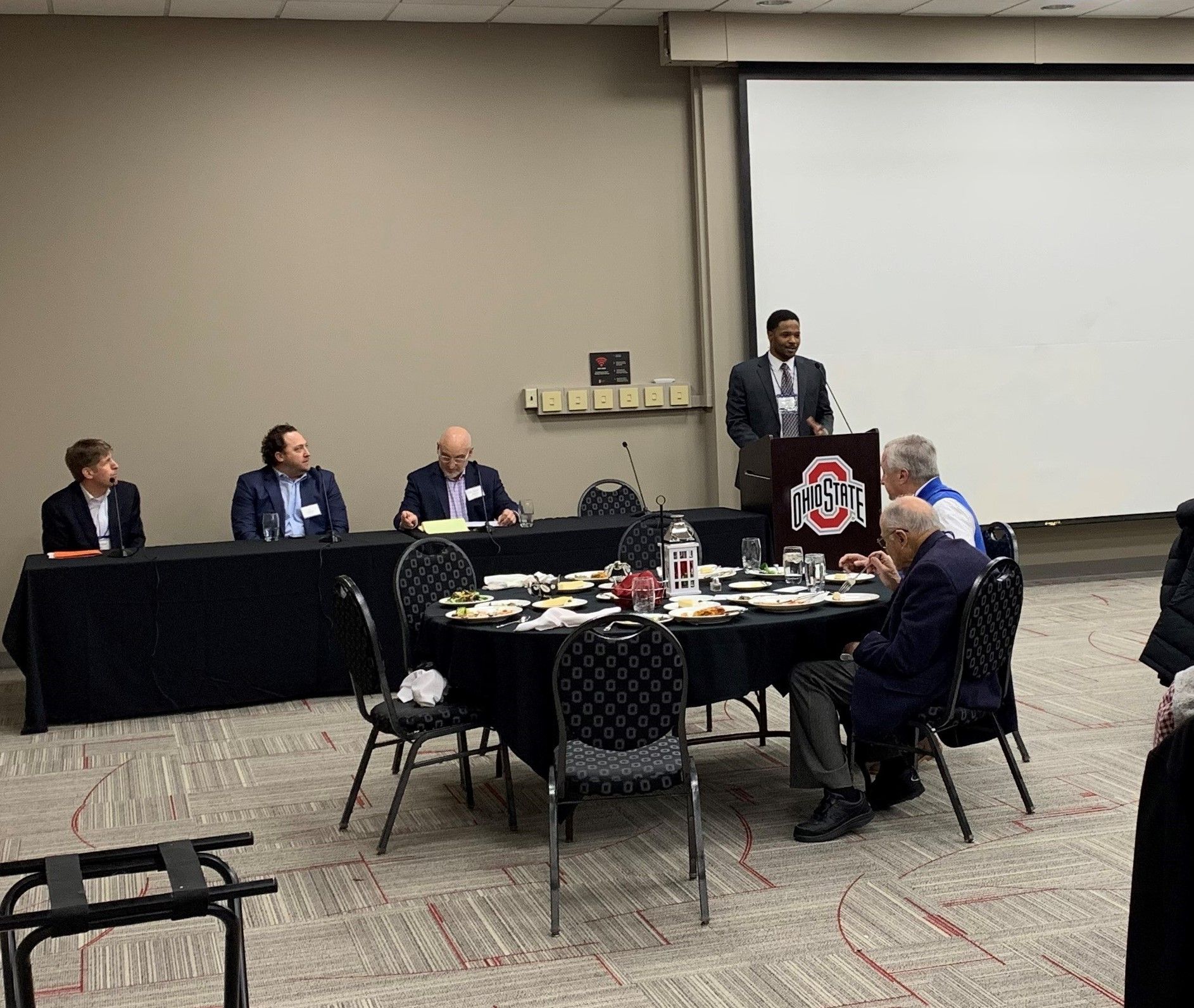 A group of people are sitting at tables in a conference room while a man stands at a podium.
