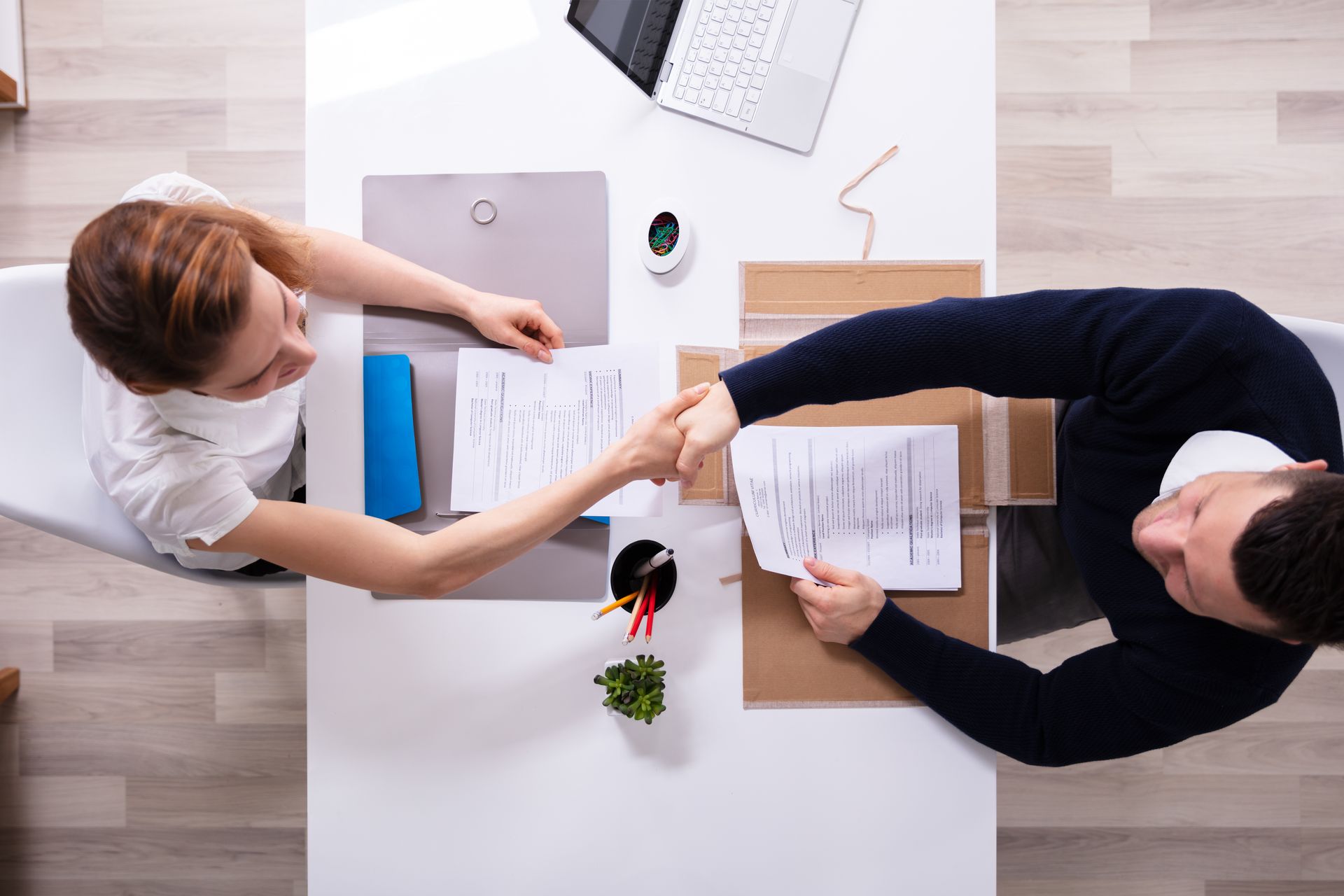 Two people reaching across a desk during a meeting, with documents, a laptop, and a small plant on the table