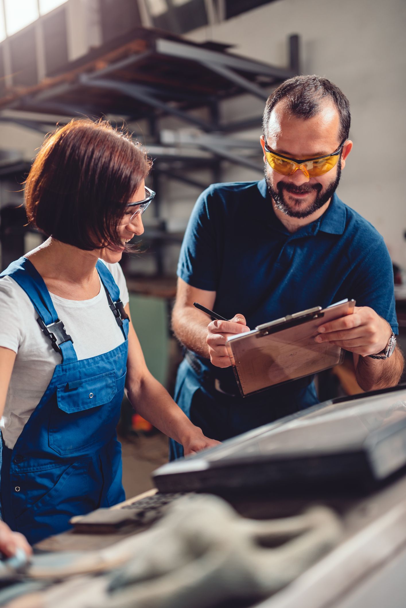 A woman and a man look at a clipboard in a workshop. The man wears safety glasses.