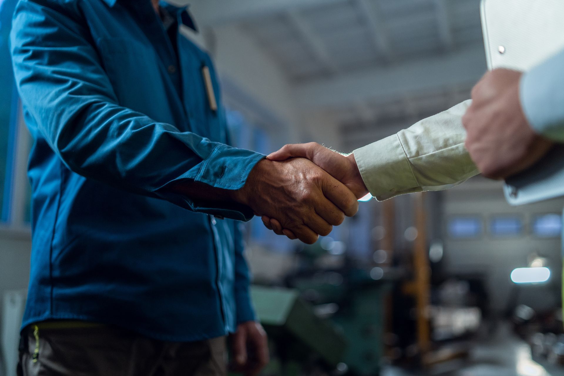 Two people shaking hands in an industrial setting, one in a blue jacket and one in a white shirt.