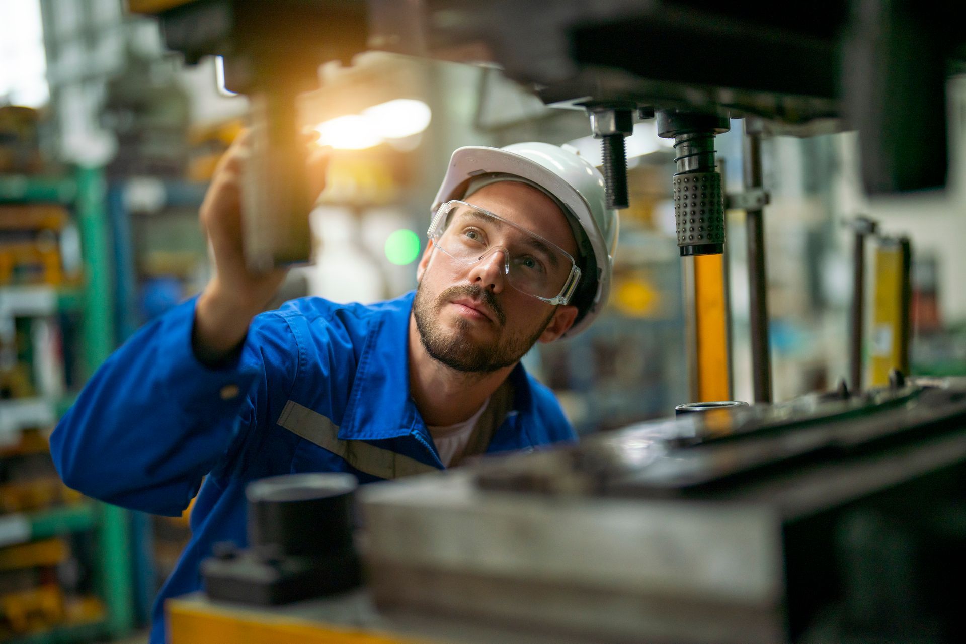Mechanic in blue overalls and hard hat inspecting machinery in a factory setting.