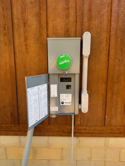 Outdoor electrical panel mounted on a wooden wall, with the panel door open revealing wiring and a label. A green sticker is on the box.