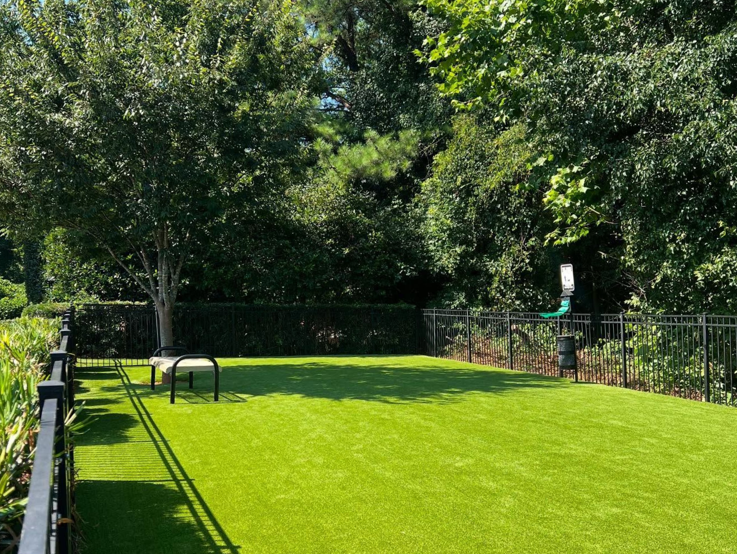 A fenced-in dog park with artificial turf, a small bench under a tree, and a waste bag station near the perimeter at Silver Collection at Waterford in Huntersville, NC.