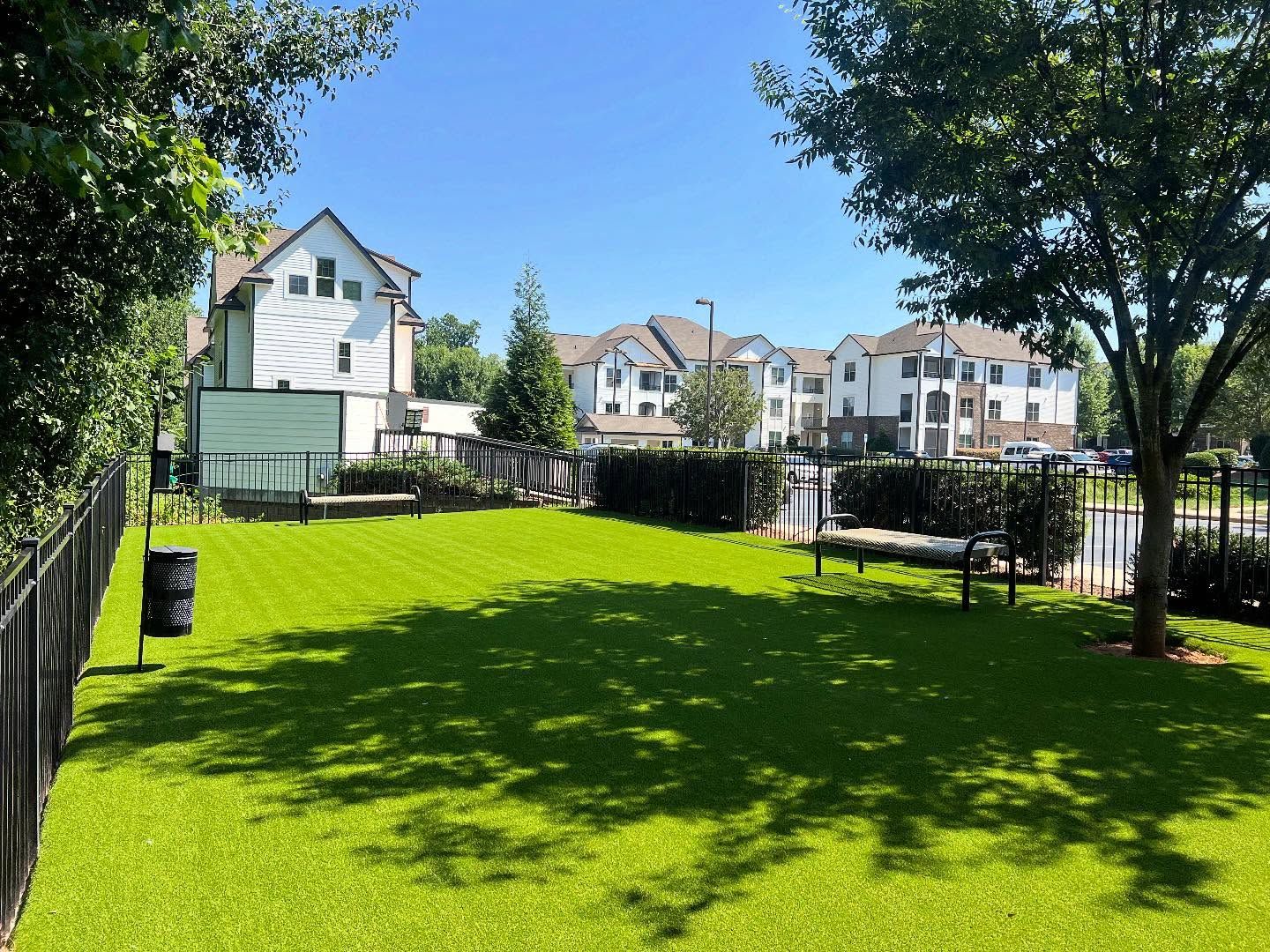 A fenced, grassy dog park area with a bench and trash bin, set against a backdrop of apartment buildings on a sunny day at Silver Collection at Waterford in Huntersville, NC.