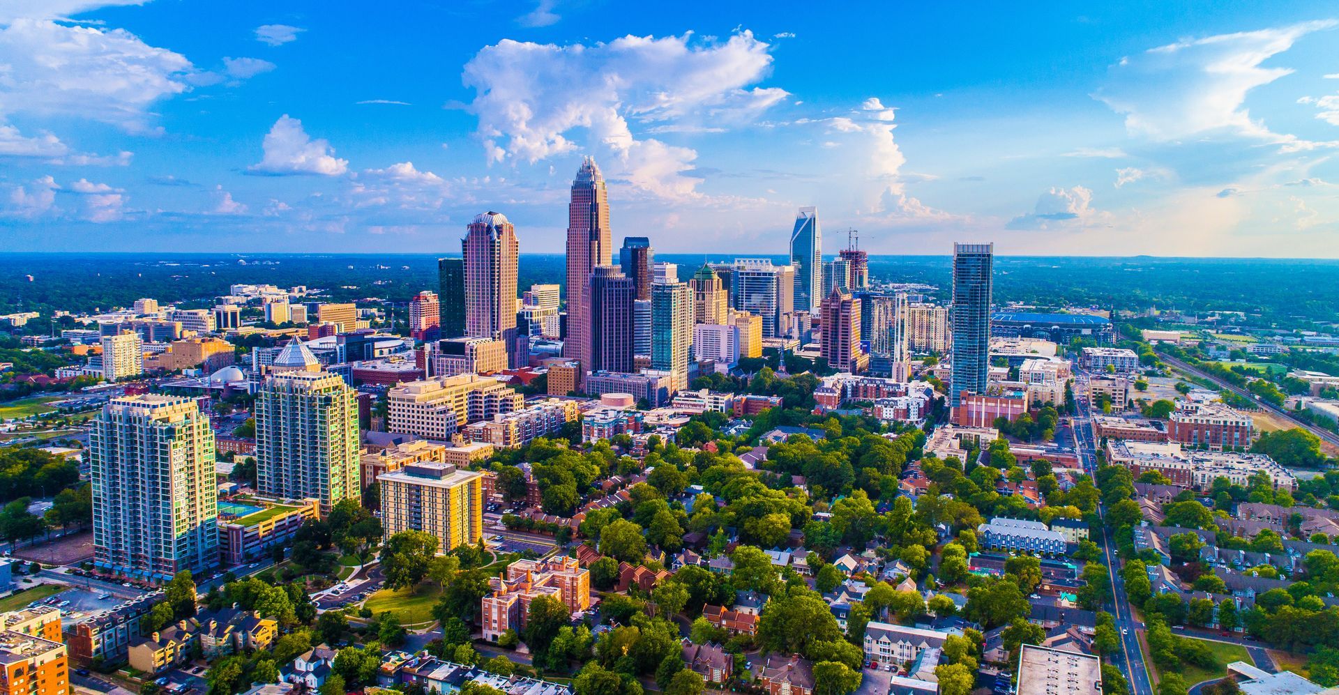 Aerial view of a modern city skyline with tall buildings, greenery, and a bright blue sky with clouds.