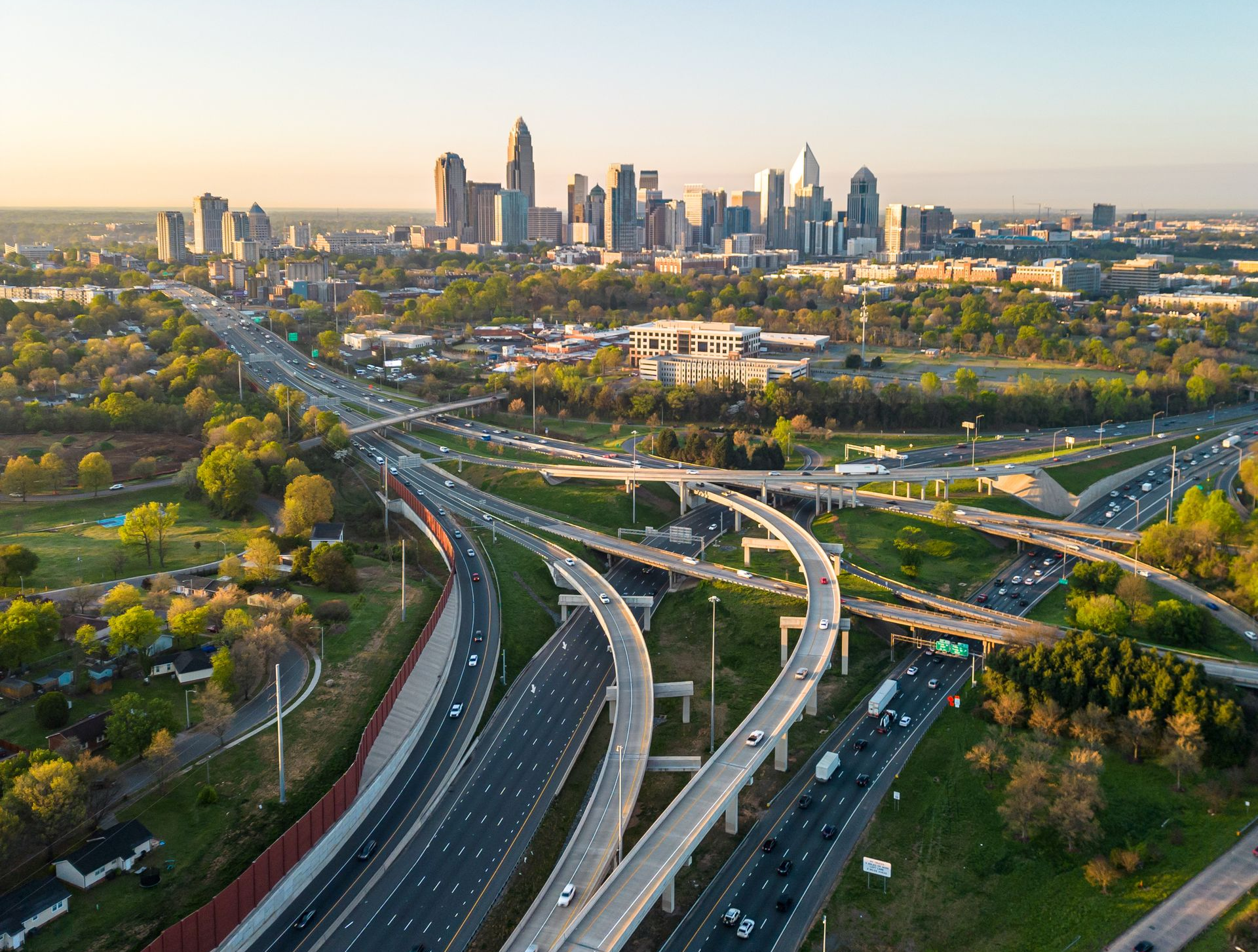 Curving highways leading toward a city skyline at sunset, with green trees and overpasses in the foreground