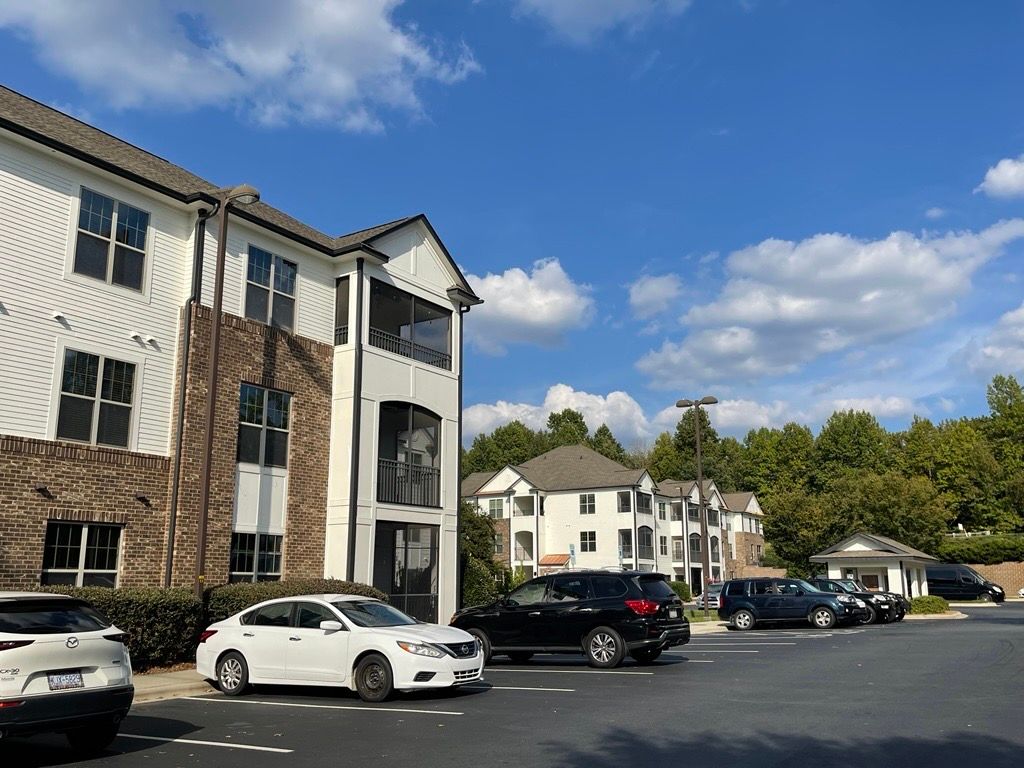 An apartment complex with white siding, brick accents, and a parking lot with several cars under a bright, cloudy sky.