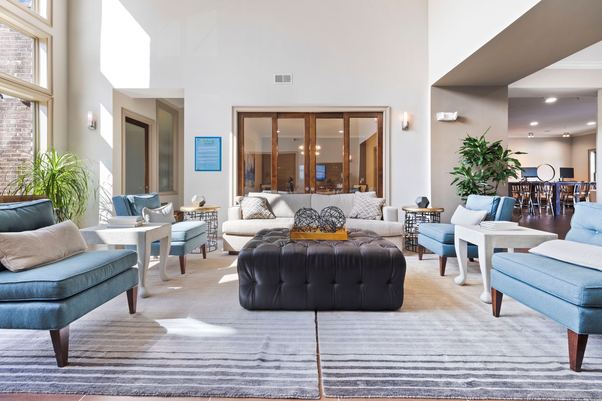 A spacious, well-lit lounge area features blue accent chairs, a central dark ottoman, and a white sofa on a large rug at Silver Collection at Waterford in Huntersville, NC.