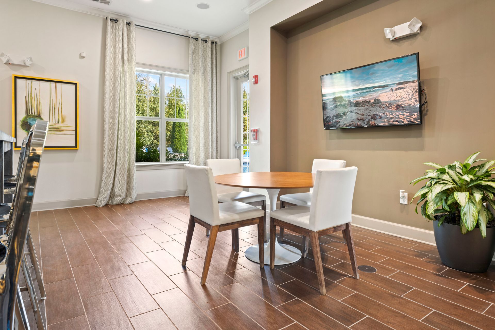 A dining area with a round wood table, four white chairs, a wall-mounted TV, a large plant, and a window with curtains at Silver Collection at Waterford in Huntersville, NC.