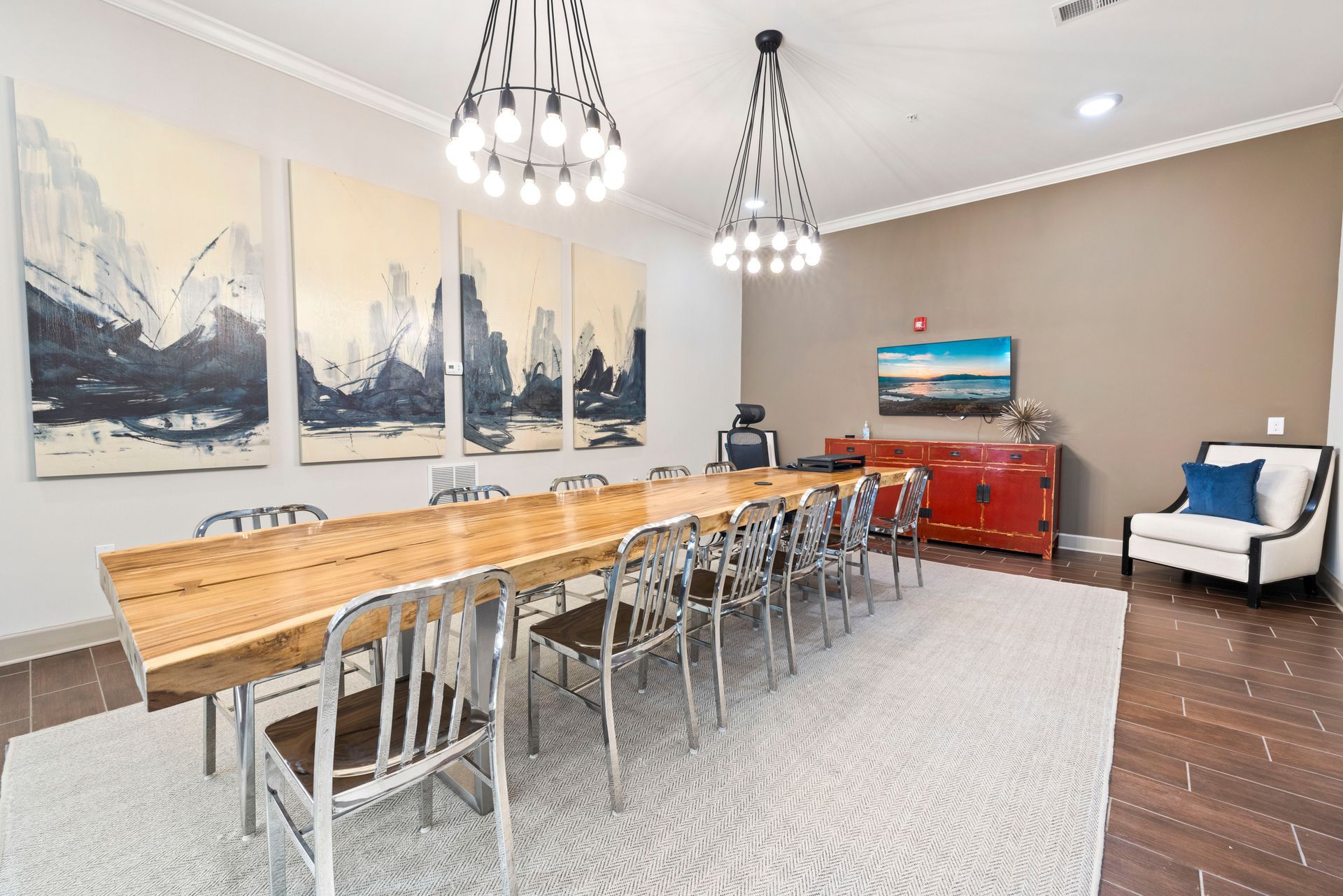 A conference room with a long wood table, metal chairs, modern chandeliers, abstract wall art, and a white armchair at Silver Collection at Waterford in Huntersville, NC.
