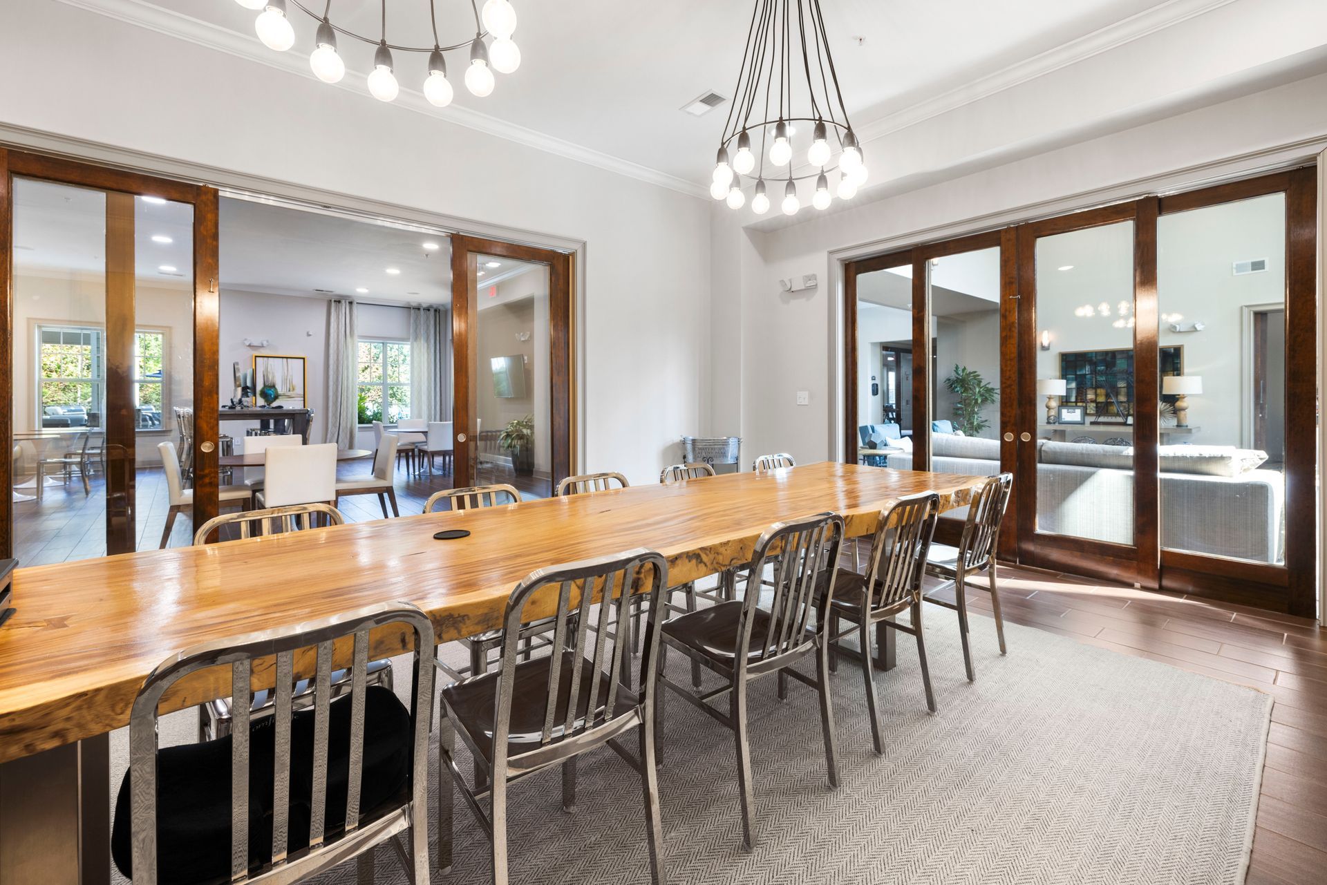 A long wooden dining table with metal chairs sits in a bright room with large glass doors and two modern chandeliers at Silver Collection at Waterford in Huntersville, NC.