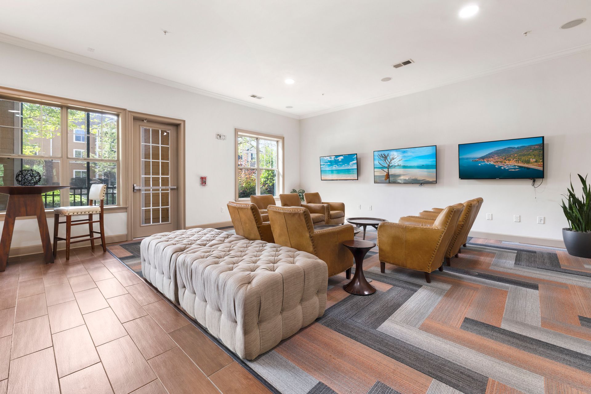 A modern lounge area with a tufted ottoman, leather armchairs, and three mounted TVs on a white wall over a patterned rug at Silver Collection at Waterford in Huntersville, NC.
