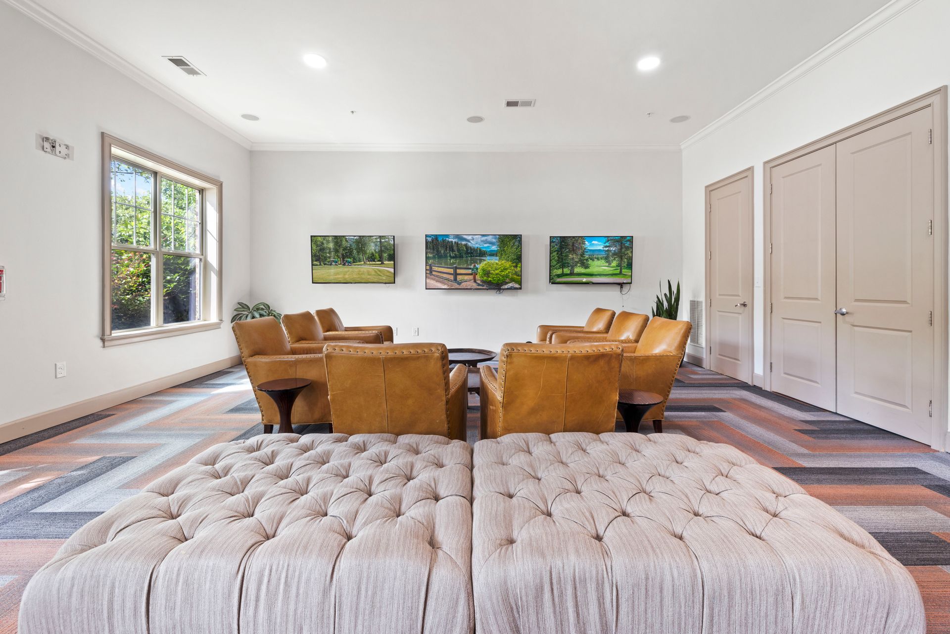 A lounge area with two large tufted ottomans in the foreground, facing leather armchairs and three wall-mounted TVs at Silver Collection at Waterford in Huntersville, NC.
