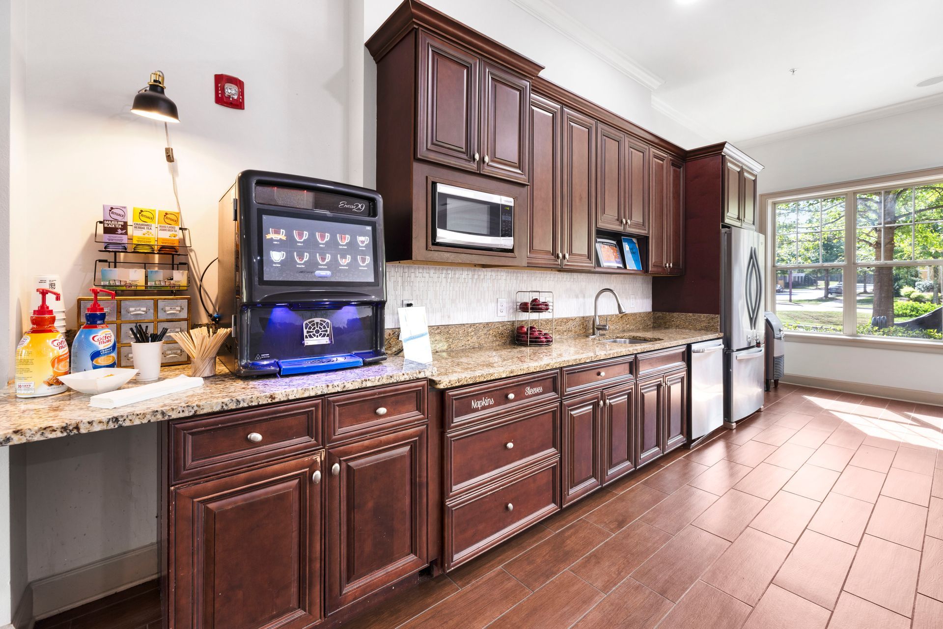 A coffee station with a touch-screen machine and snacks, set on a granite countertop with dark cabinets in a kitchen area at Silver Collection at Waterford in Huntersville, NC.