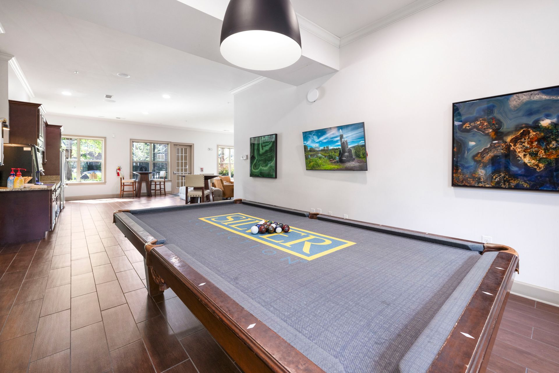 A pool table with grey felt sits in a bright, modern open-concept room with dark wood floors and art on the wall at Silver Collection at Waterford in Huntersville, NC.