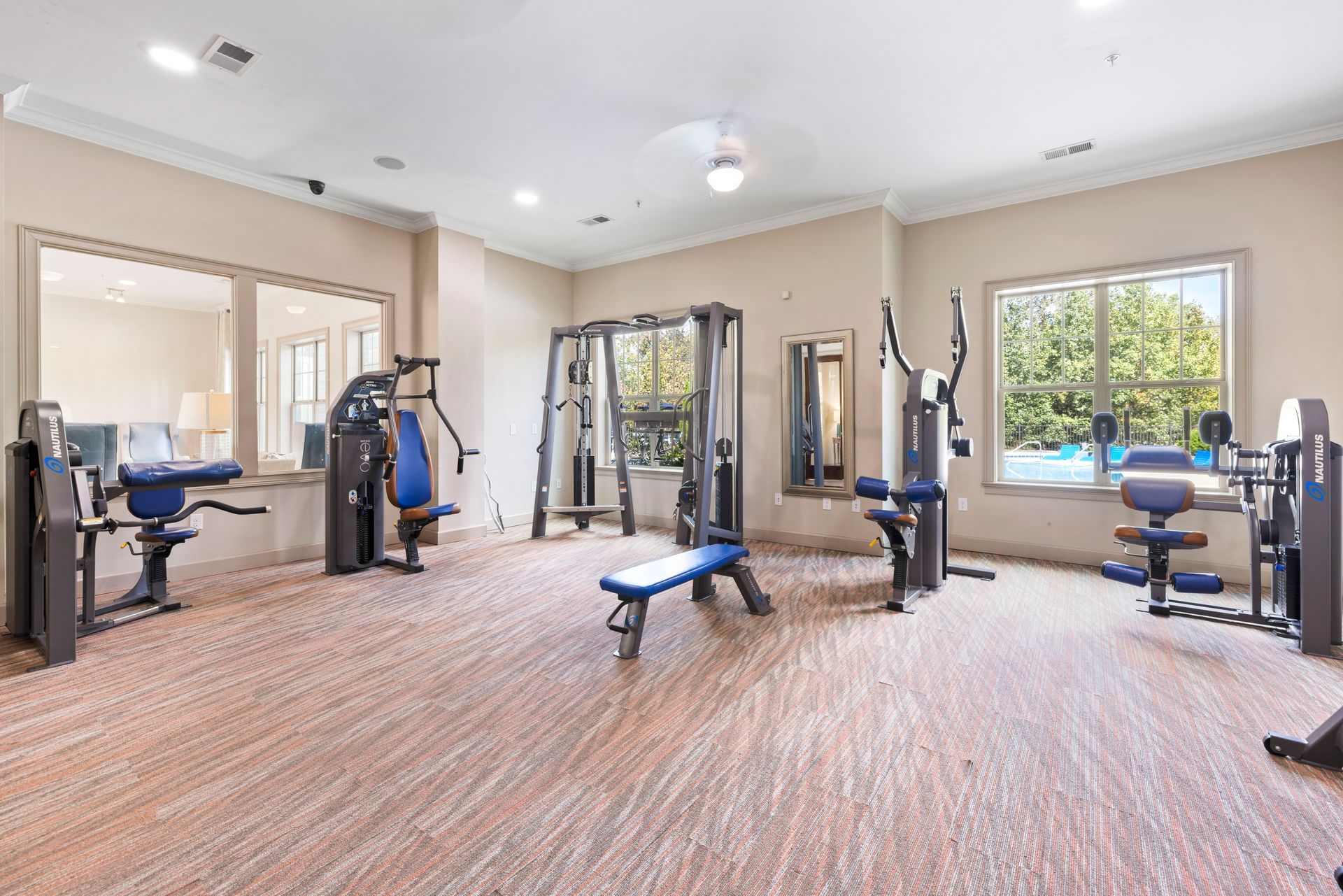 A well-lit, empty apartment fitness center with various weight machines, a workout bench, and large windows at Silver Collection at Waterford in Huntersville, NC.