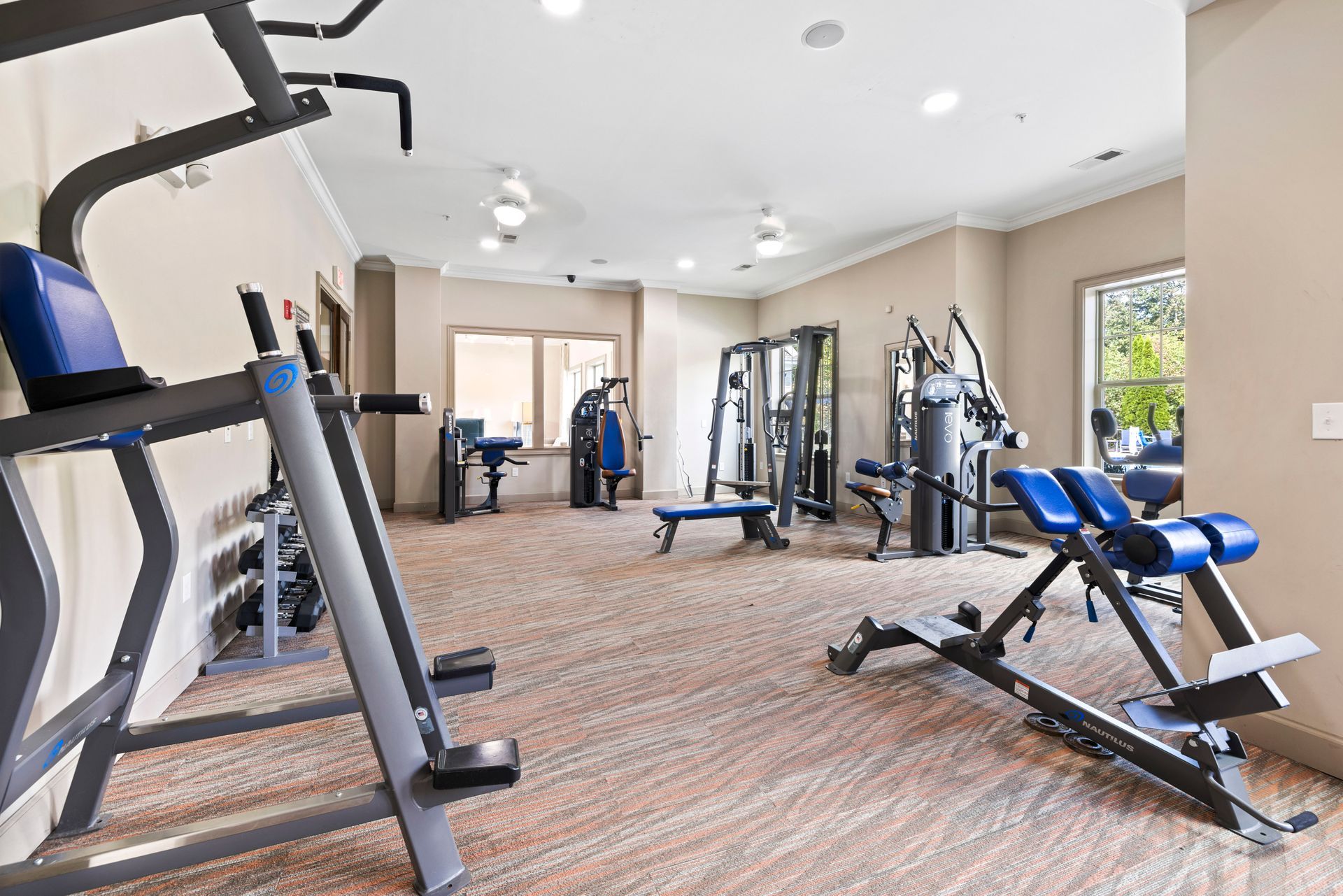 A bright, neutral-toned fitness center featuring multiple strength training machines, blue padding, and carpeted floors at Silver Collection at Waterford in Huntersville, NC.