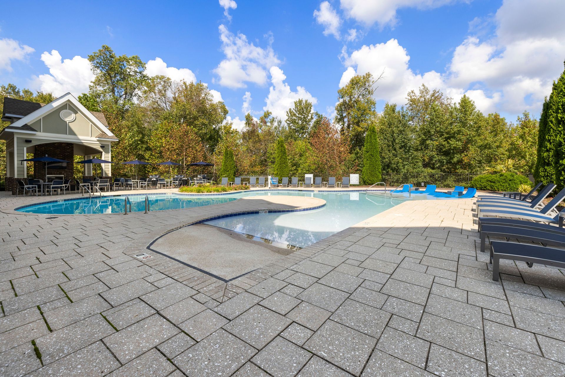 A swimming pool with a shallow sun shelf and adjacent pavilion, surrounded by trees under a bright blue, cloudy sky at Silver Collection at Waterford in Huntersville, NC.