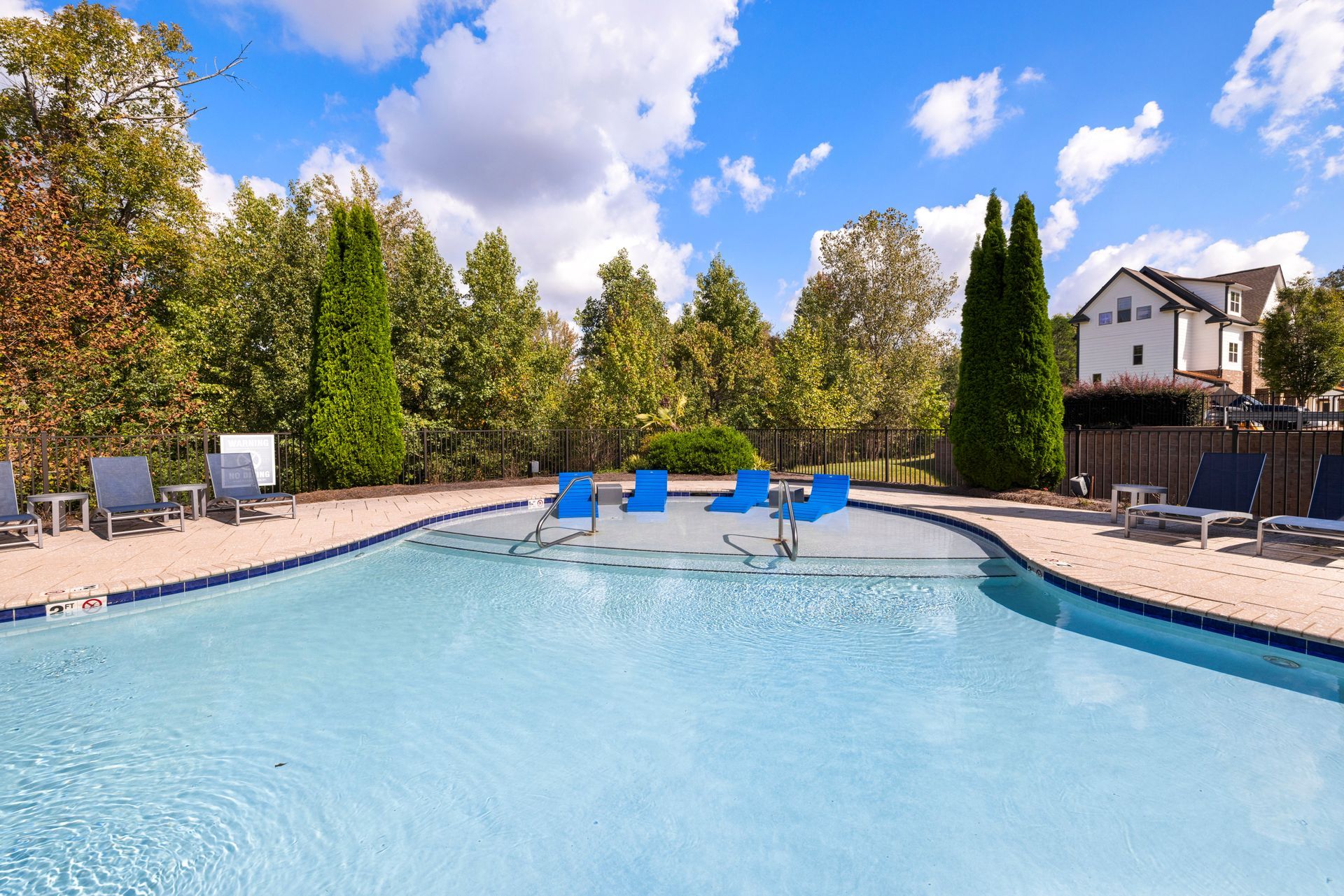 An outdoor swimming pool with bright blue water, surrounded by tan patio furniture and lush green trees under a blue sky at Silver Collection at Waterford in Huntersville, NC.