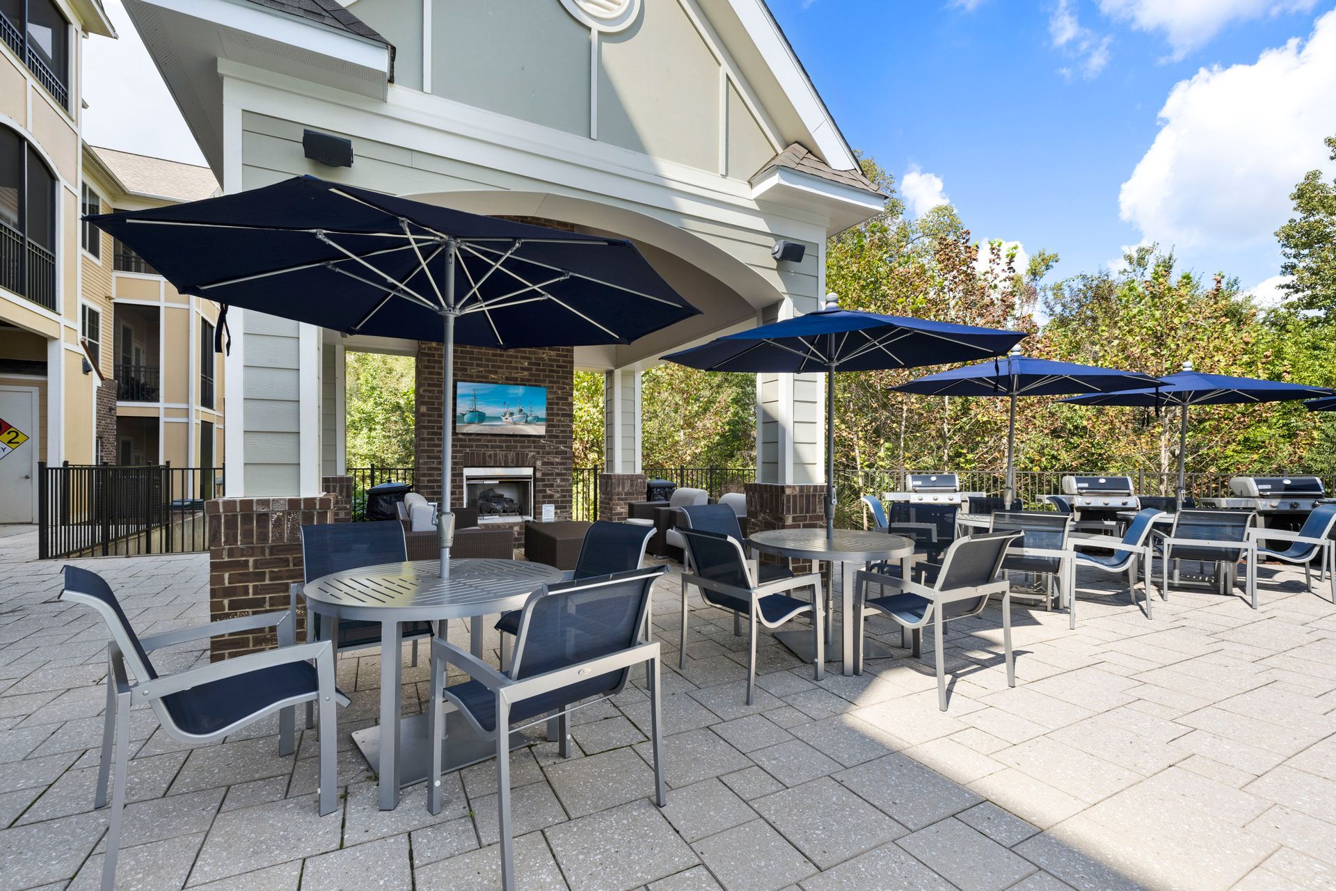 An outdoor patio with tables, chairs, and blue umbrellas, featuring a covered lounge area with a fireplace and TV at Silver Collection at Waterford in Huntersville, NC.