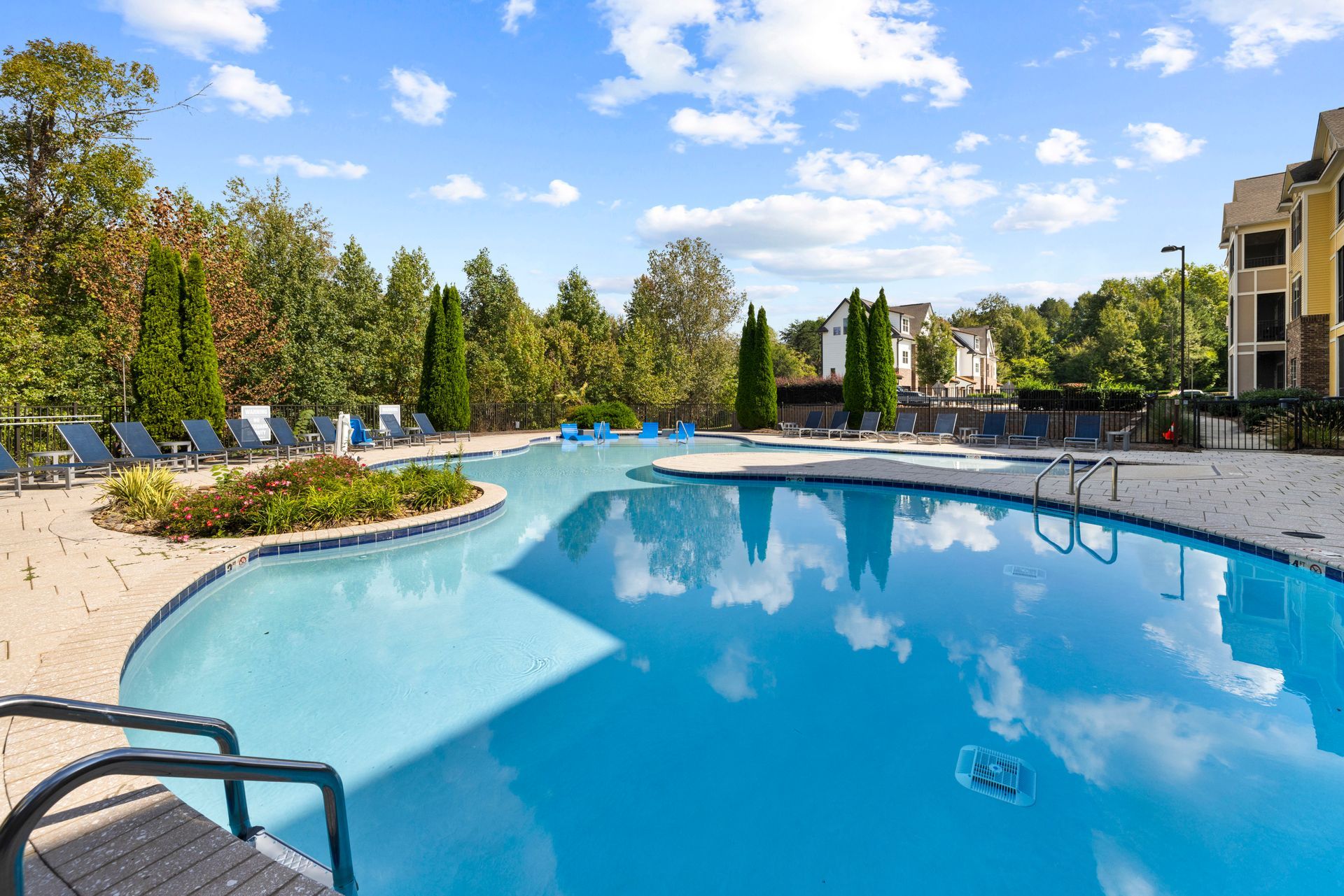 An outdoor swimming pool surrounded by a patio, landscaping, and apartment buildings under a sunny blue sky at Silver Collection at Waterford in Huntersville, NC.
