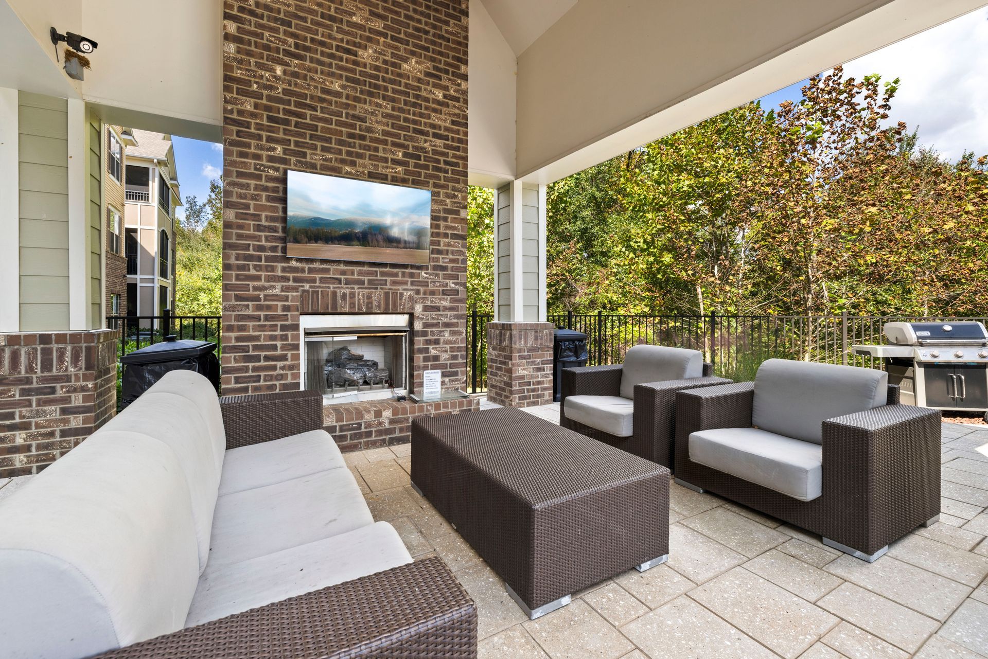 A covered patio featuring a brick fireplace with a mounted television, wicker furniture with cushions, and a grill area at Silver Collection at Waterford in Huntersville, NC.