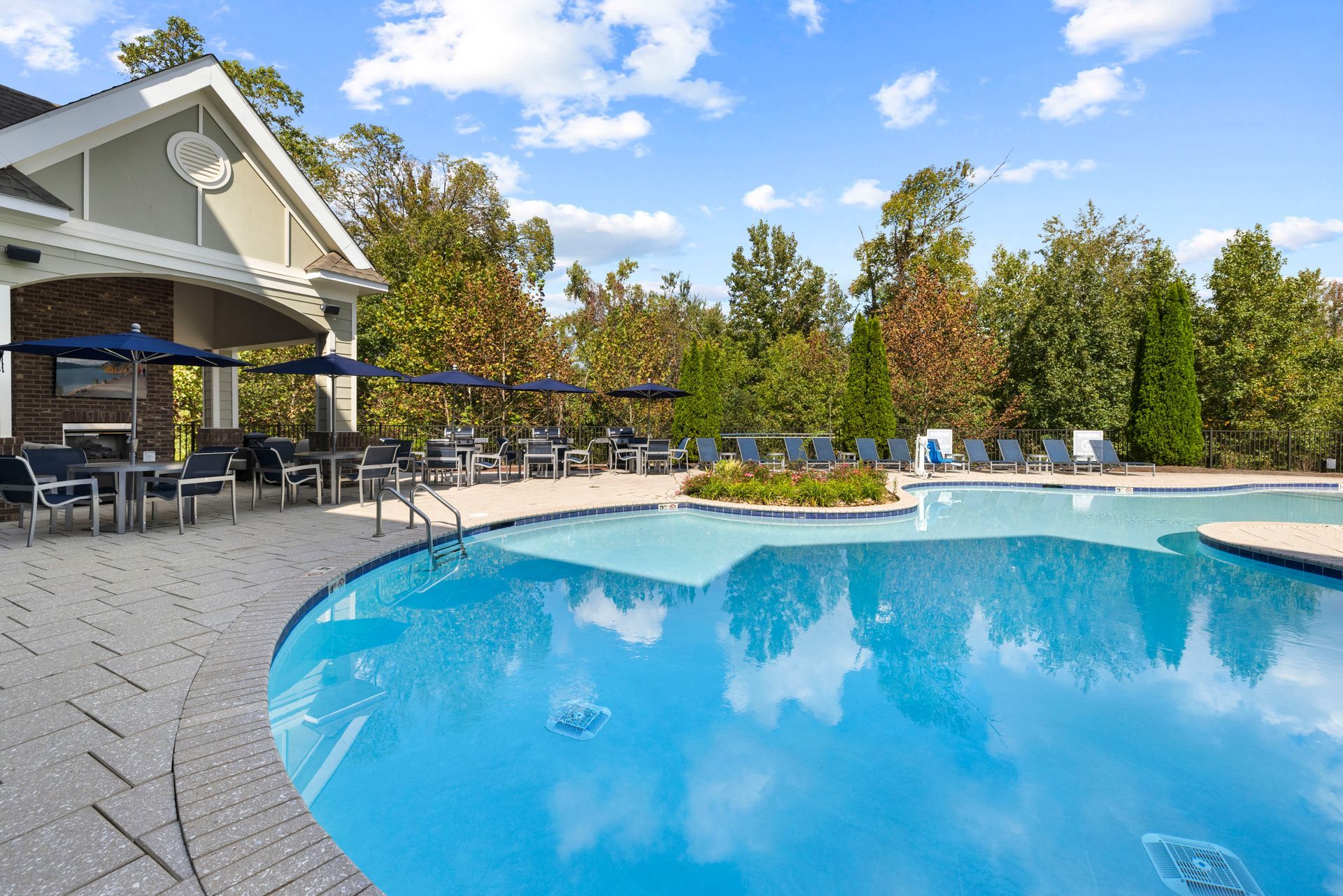 A bright outdoor swimming pool with a stone patio, dining tables, umbrellas, and surrounding trees under a sunny sky at Silver Collection at Waterford in Huntersville, NC.