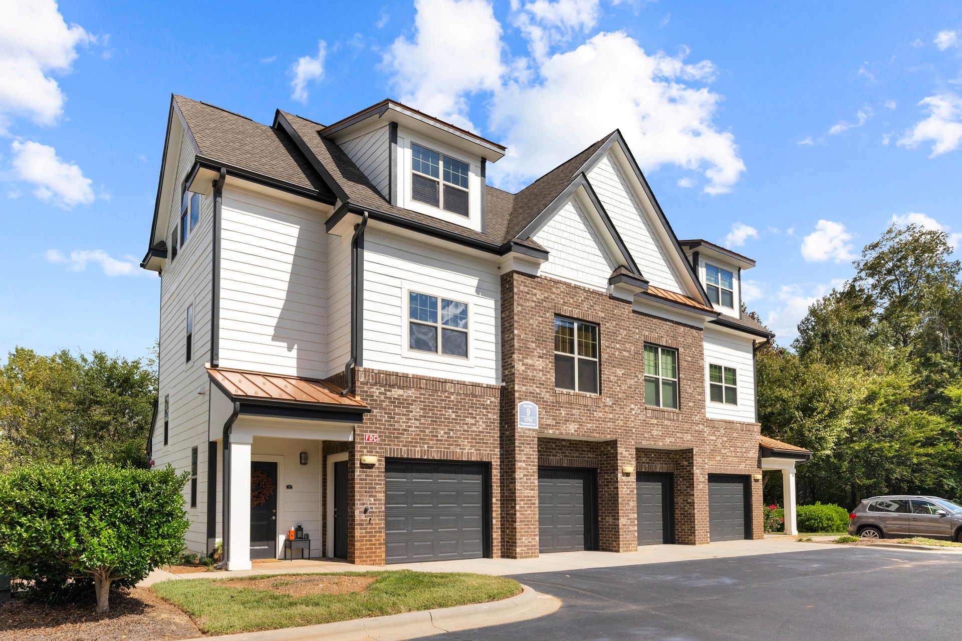 A three-story townhome with a white upper exterior, brick lower level, and four garage doors under a blue, cloudy sky at Silver Collection at Waterford in Huntersville, NC.
