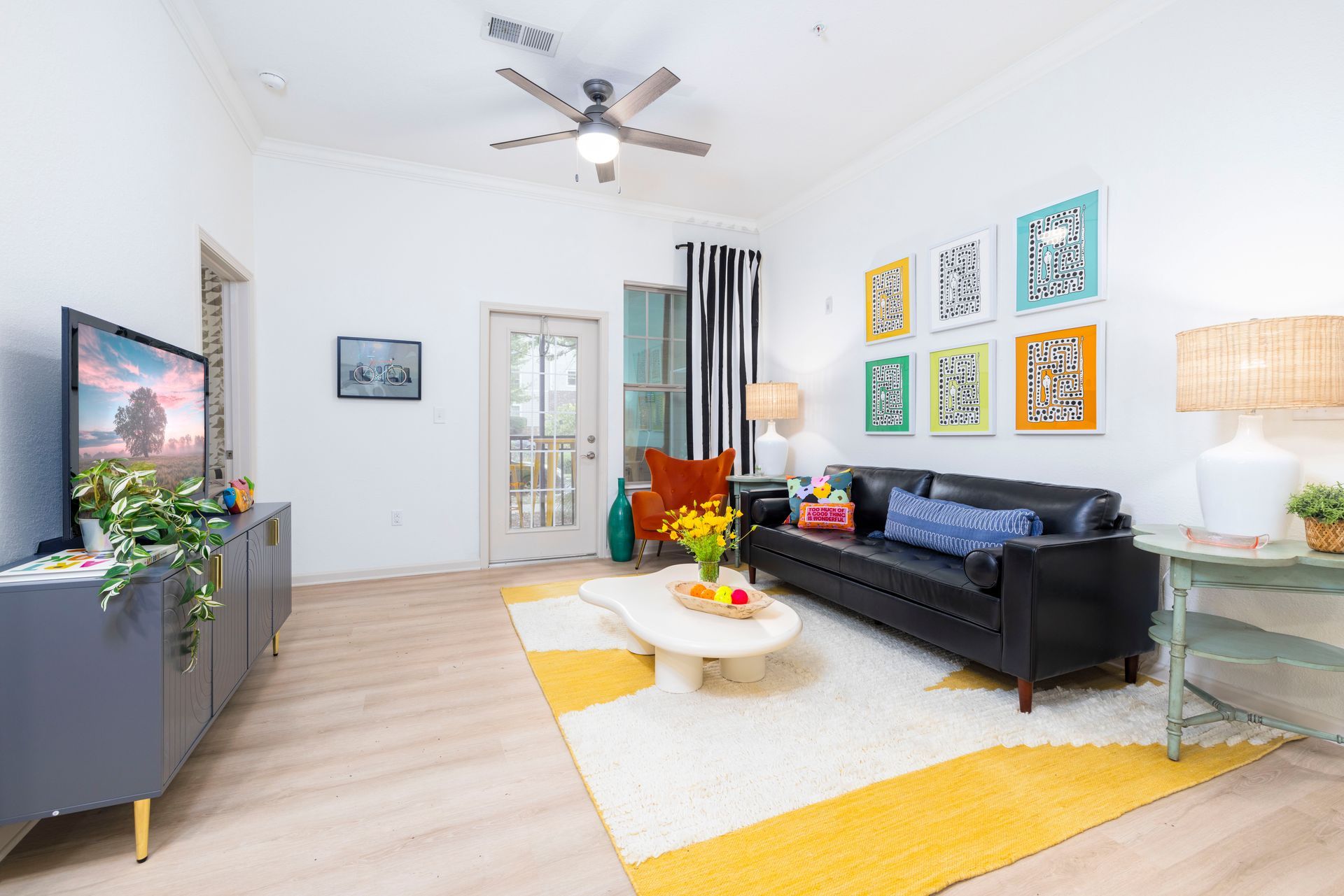 A bright, modern living room with a black sofa, a patterned rug, a light-colored coffee table, and colorful wall art.