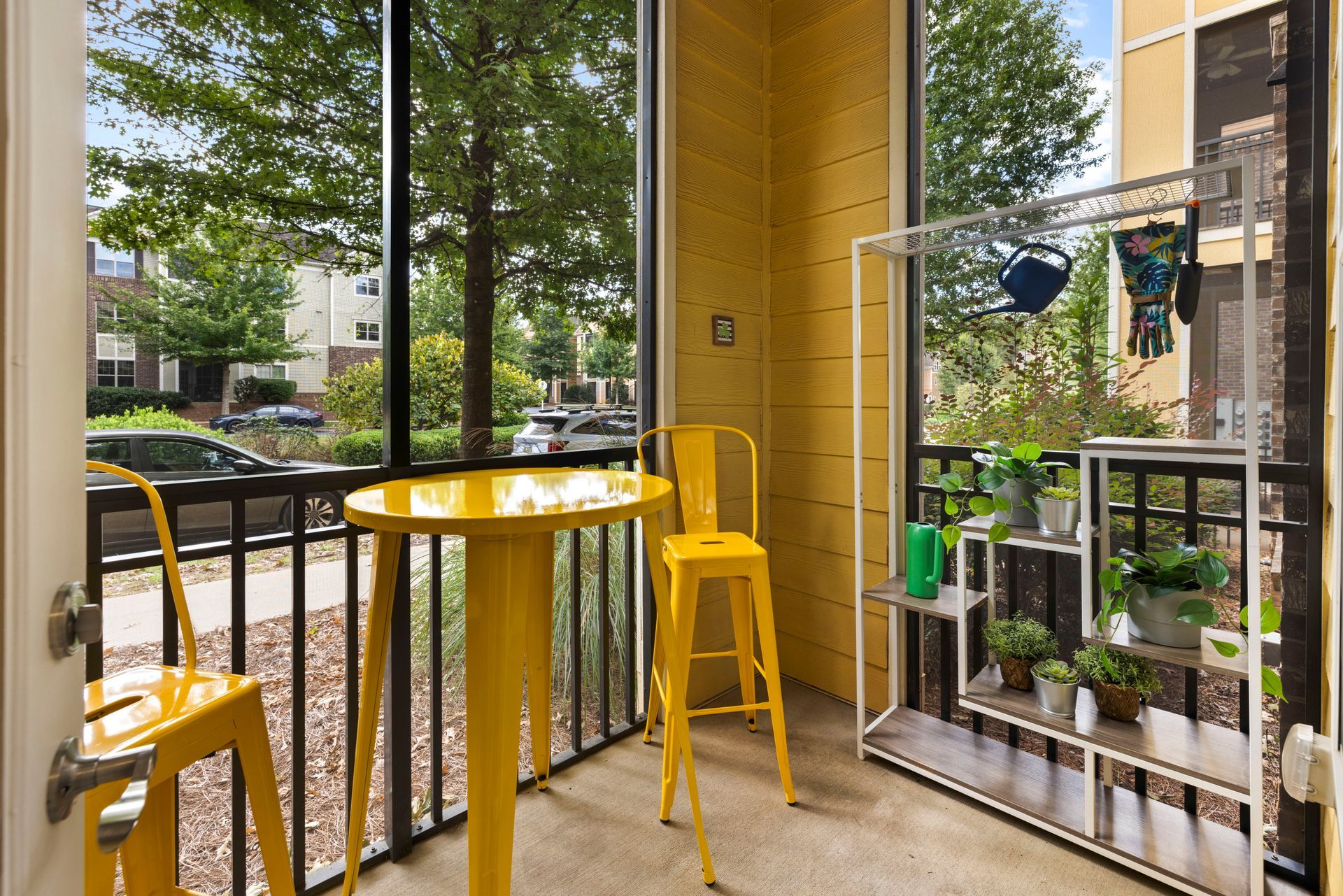 A small, sunny balcony with a round yellow bistro table, two yellow stools, and a metal shelf filled with potted plants.