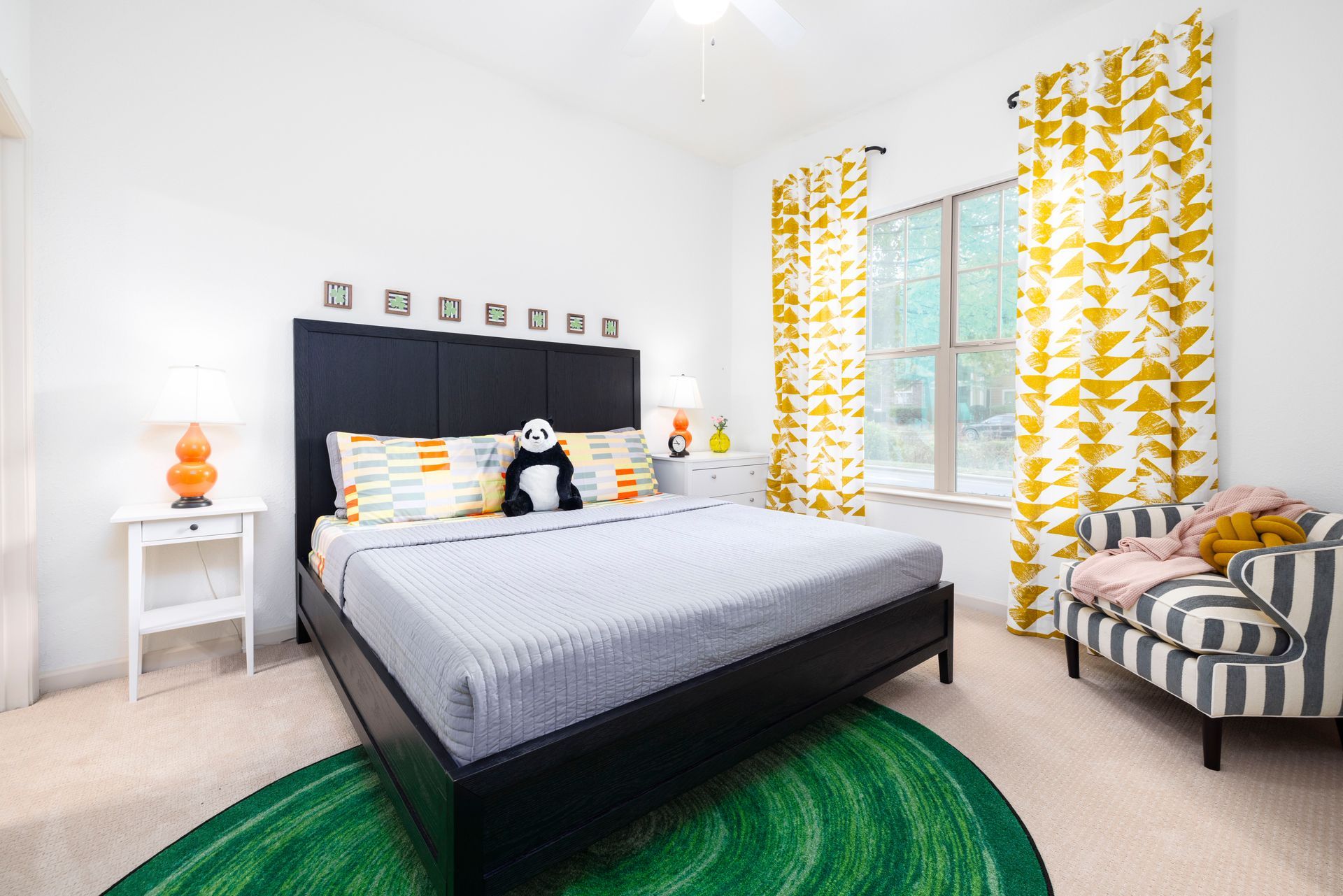 A bedroom with a black bed frame, grey bedding, a green circular rug, yellow patterned curtains, and a striped chair.