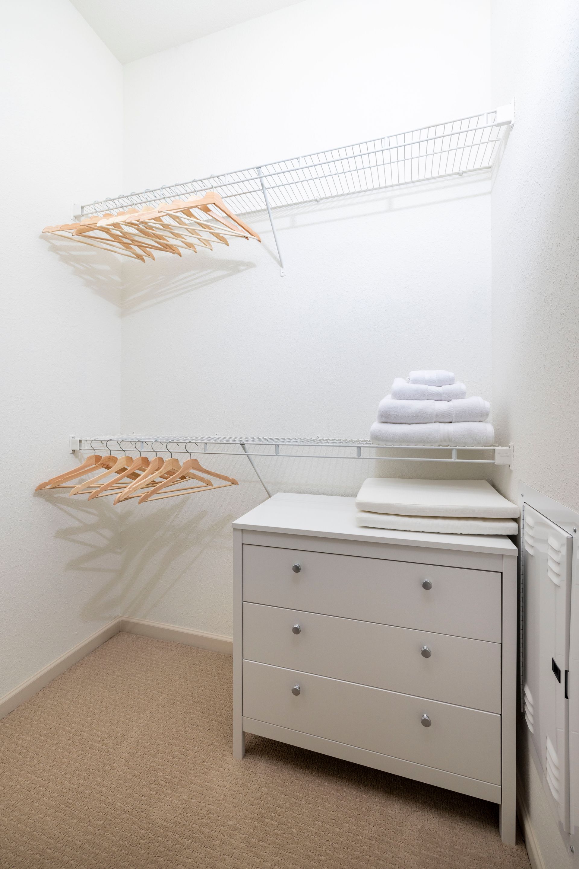 A white dresser with stacked towels sits in a walk-in closet with wire shelving and wooden hangers.