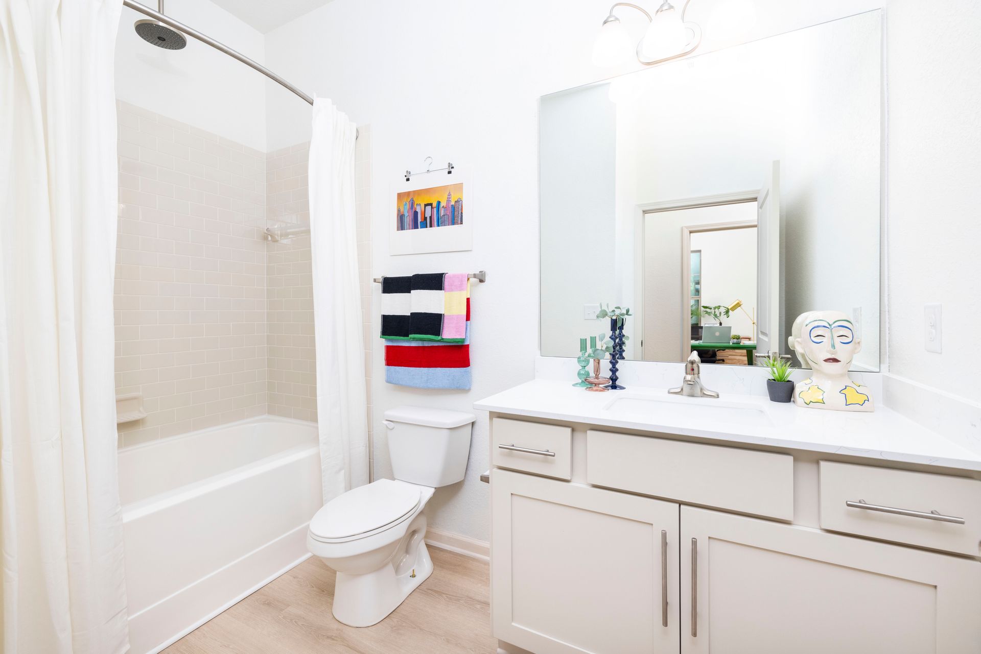 A bright, white bathroom with a shower-tub combo, a toilet, a vanity with a large mirror, and colorful hanging towels at Silver Collection at Waterford in Huntersville, NC.