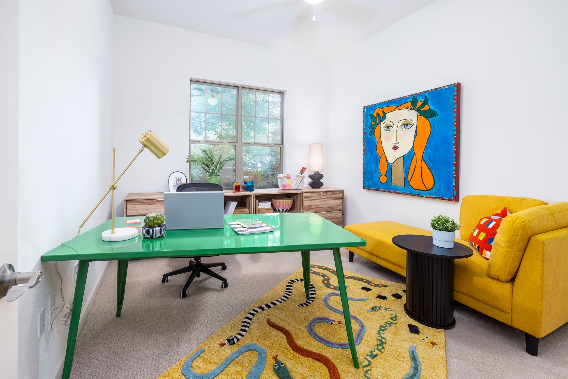 A bright home office with a green desk, a yellow sofa, patterned rug, and large abstract art piece on a white wall.