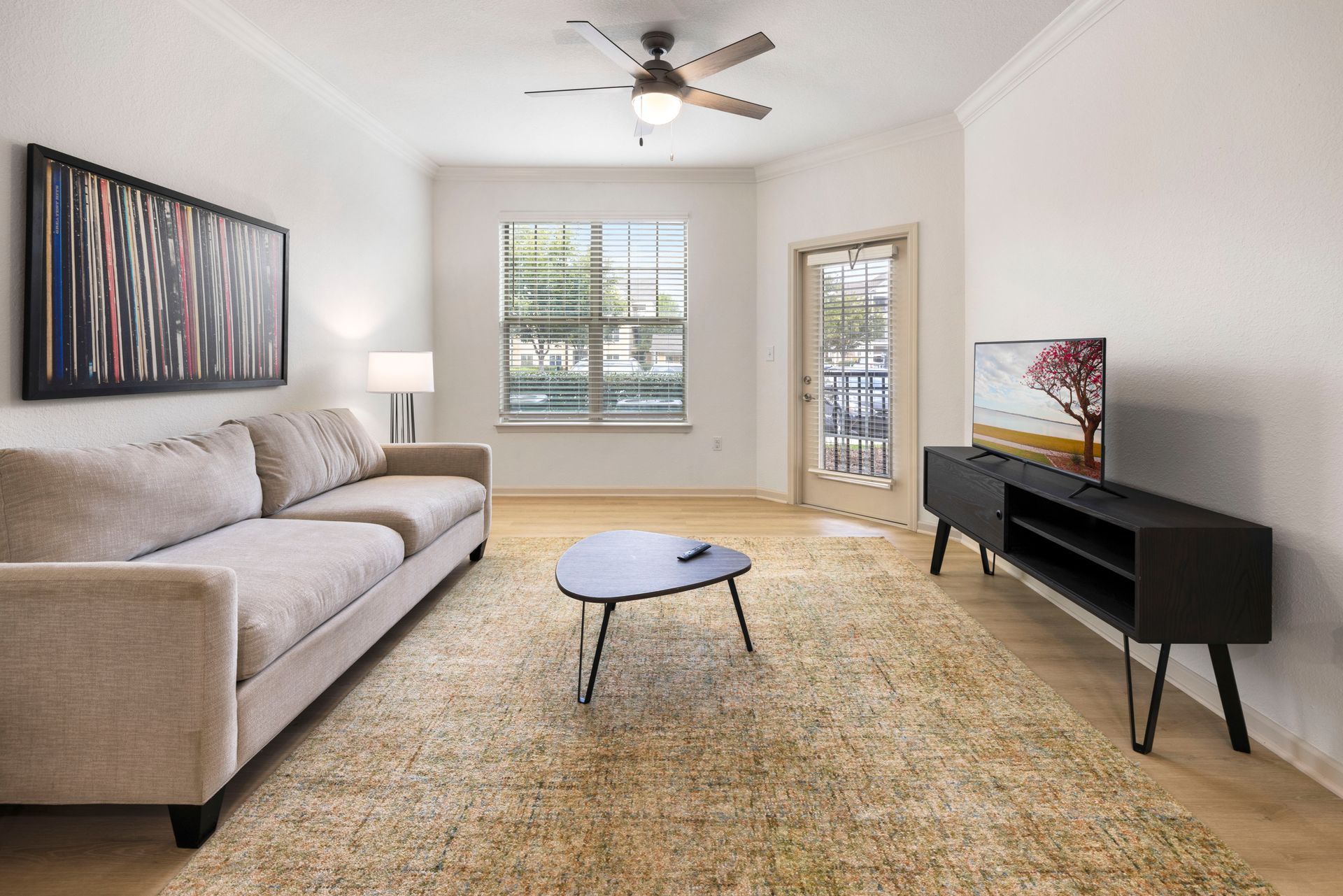 A furnished living room with a neutral sofa, patterned wall art, a coffee table, a TV on a black stand, and a ceiling fan at Silver Collection at Waterford in Huntersville, NC.
