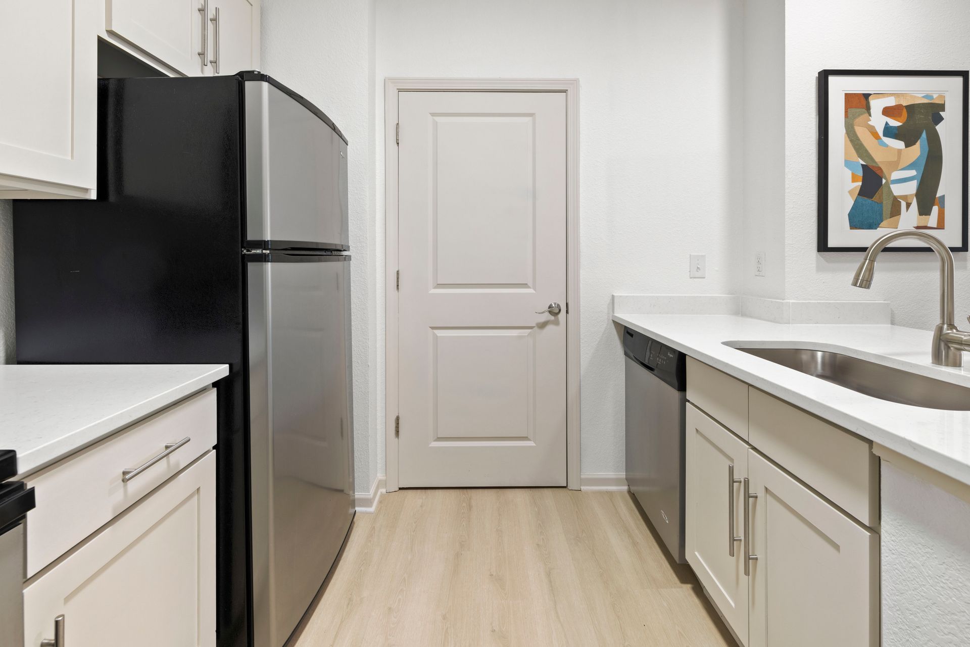 Modern kitchen with stainless steel appliances, white cabinets, white countertops, light wood flooring, and a closed door at Silver Collection at Waterford in Huntersville, NC.